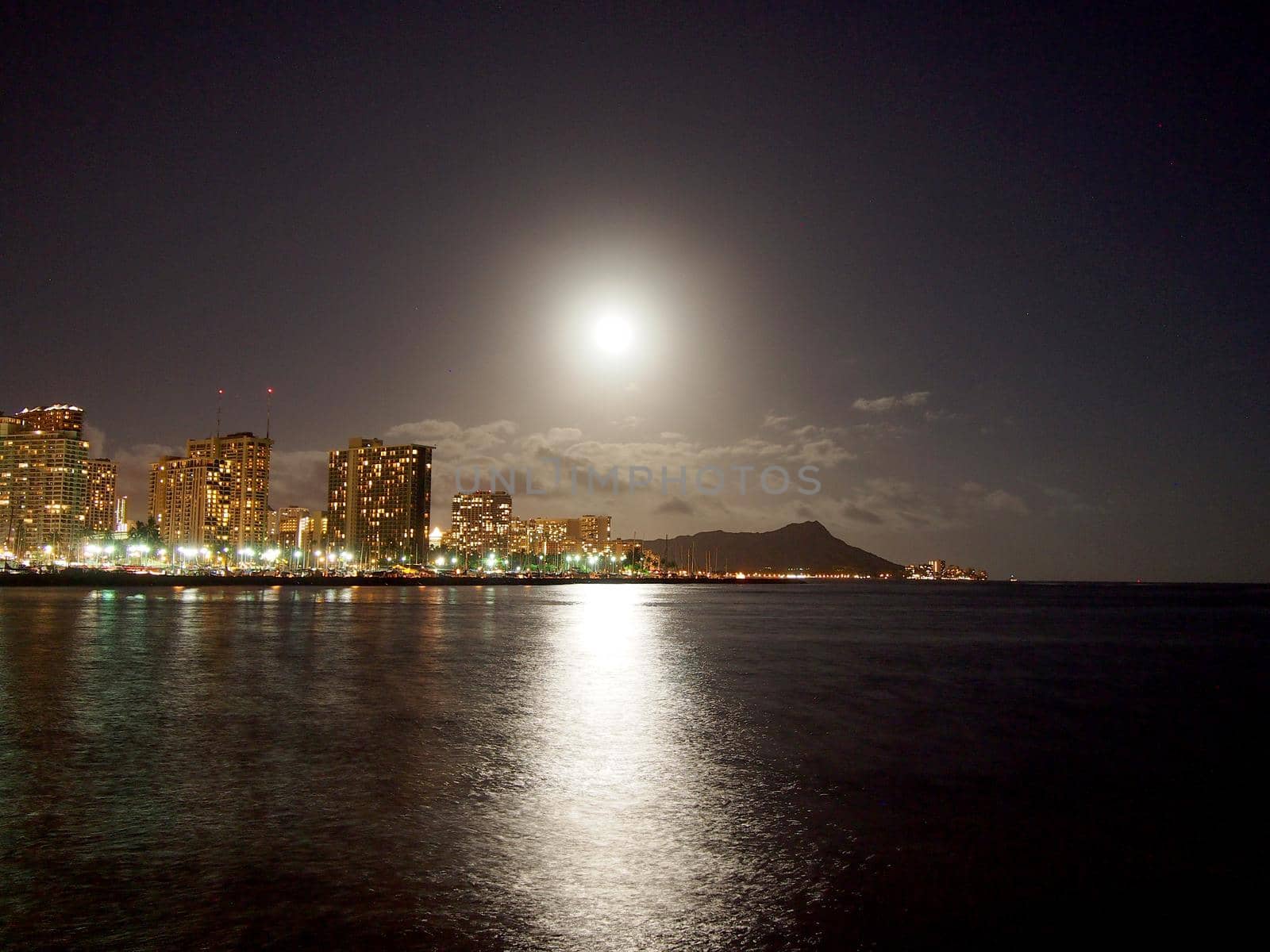 Full Large Moon hangs over Diamond Head Crater, Waikiki hotels, and Marina while reflecting on the water at Night on Oahu, Hawaii.
