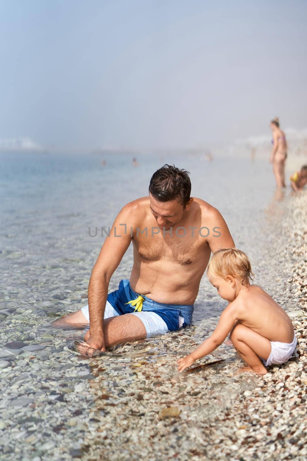Dad and a little girl are sitting in a water and poking around in the pebbles by Nadtochiy