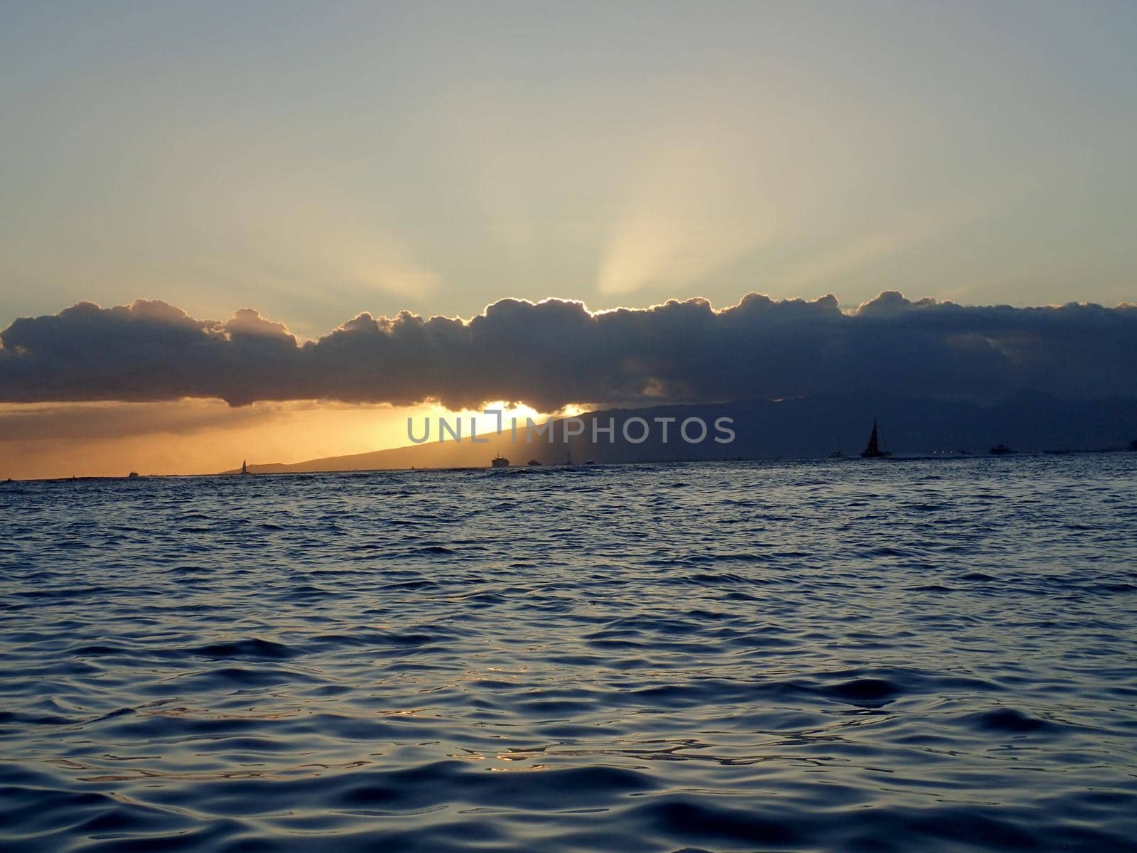Sunsets through the clouds over Waianae mountains with light reflecting on ocean and illuminating the sky with boats sailing on the water off Waikiki on Oahu, Hawaii.