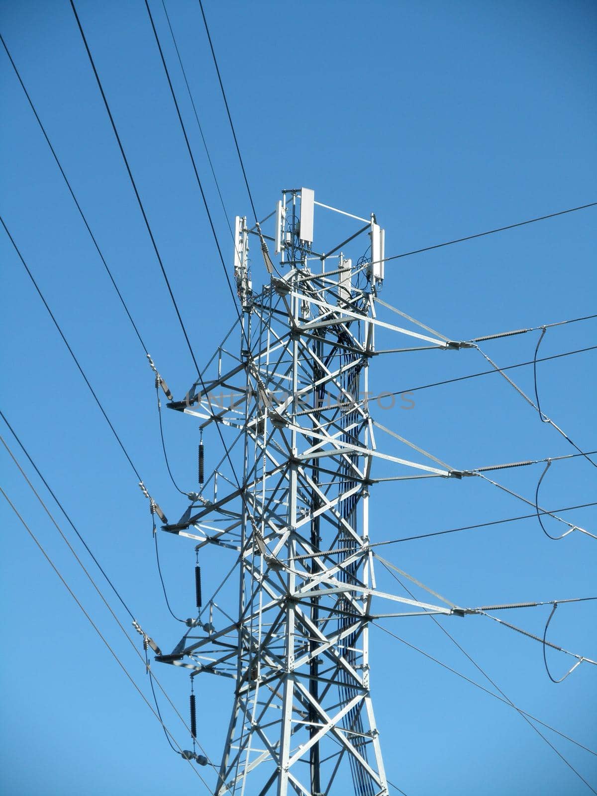 High Voltage Power Lines intersect at a large metal Utility pole with cell tower against a blue sky in San Jose, California.