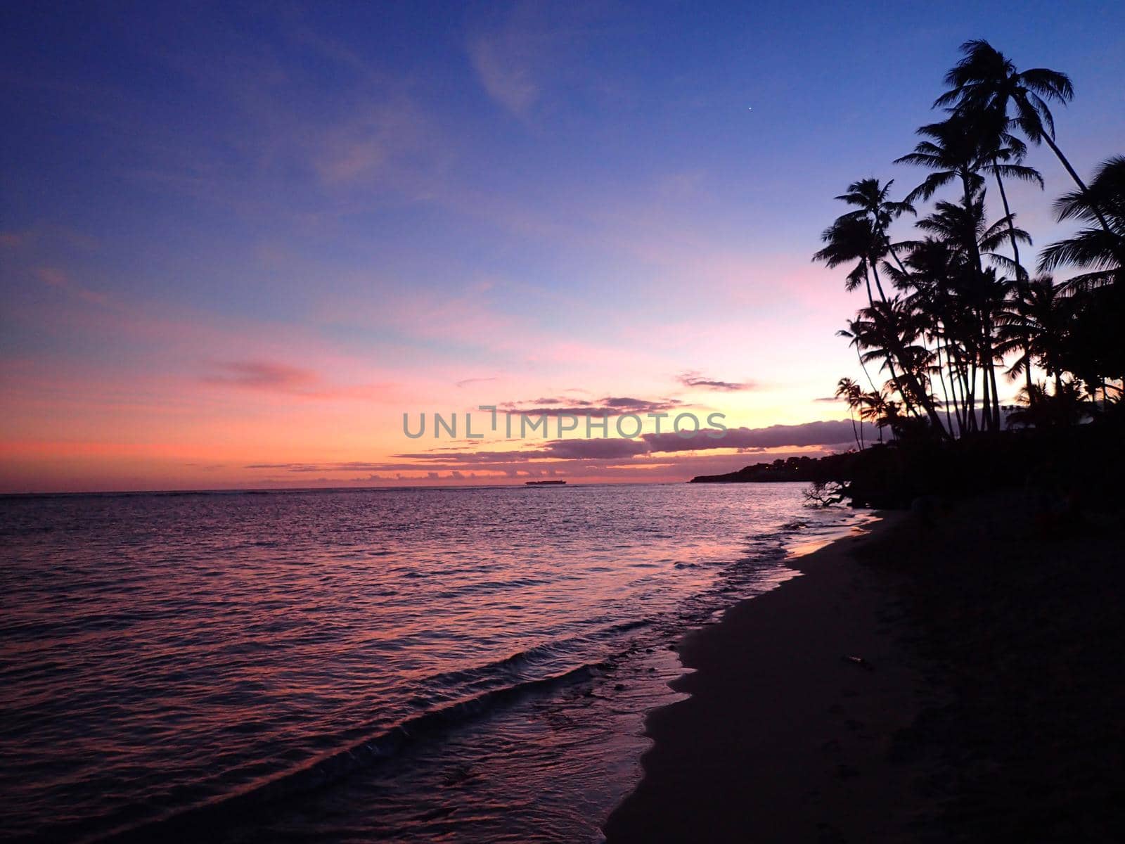 Sandy shoreline of Kahala Beach and the southern coastline at dusk with pink and orange light filling the air of Oahu, Hawaii.