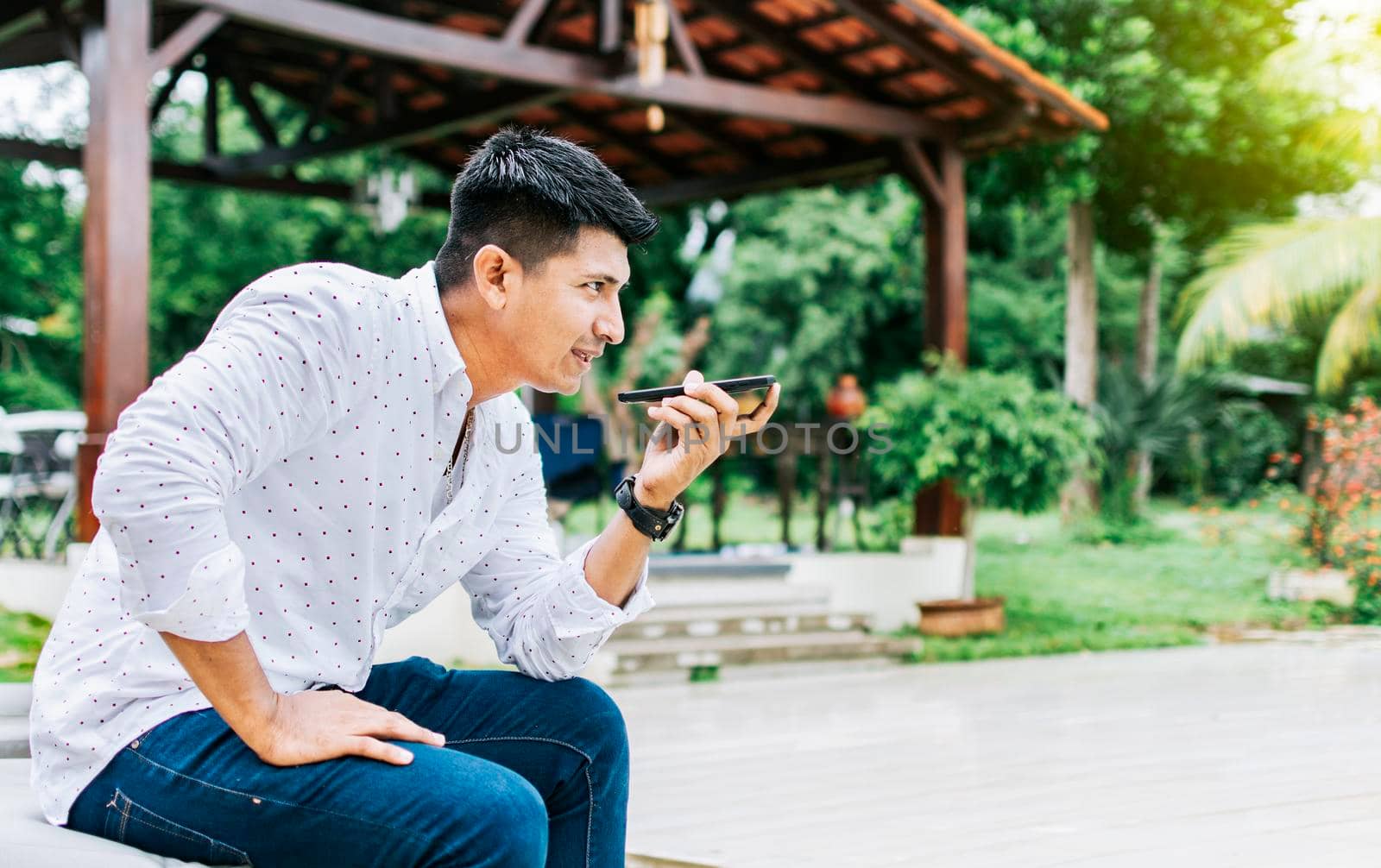 Handsome man sitting outside sending a voice message with his cell phone, Teenage sitting calling on the phone outside, Attractive man sitting sending a voice message with his phone and gesturing by isaiphoto