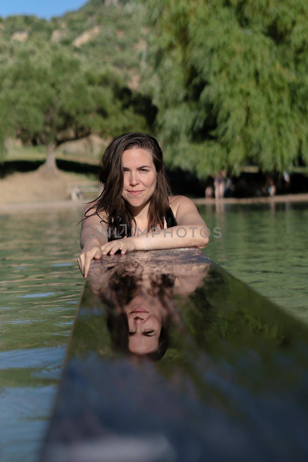 attractive young girl reflected in the water of a lake leaning on a wooden board long-haired brunette in black swimsuit