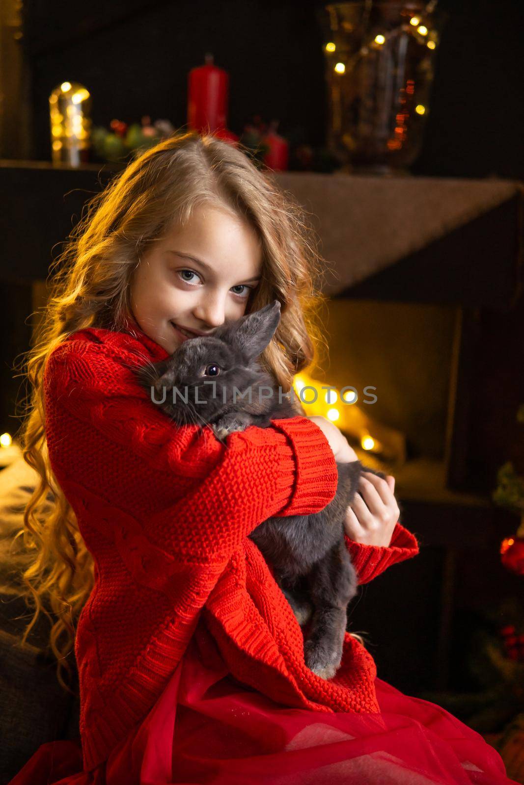 A little blonde girl with a gray rabbit in her arms next to a Christmas tree decorated with garlands, glass balls and Christmas toys and a fireplace. The concept of winter holidays is Christmas and New Year holidays. Magical festive atmosphere.