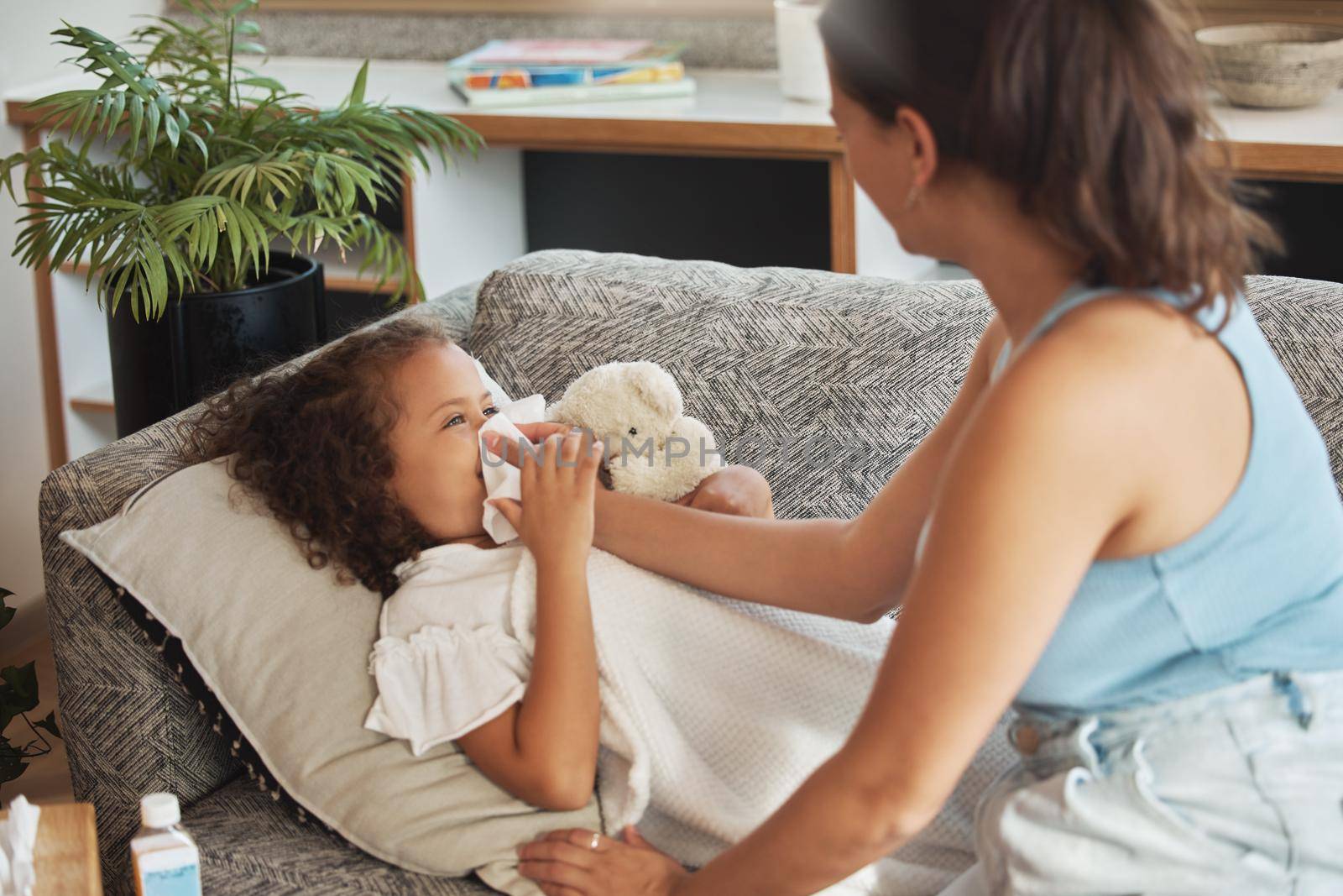 Caring, loving and worried parent taking care of ill kid. Suffering from cold, flu or covid virus. Daughter, child and sick girl lying on sofa while mother helps her blow her nose in home living room by YuriArcurs