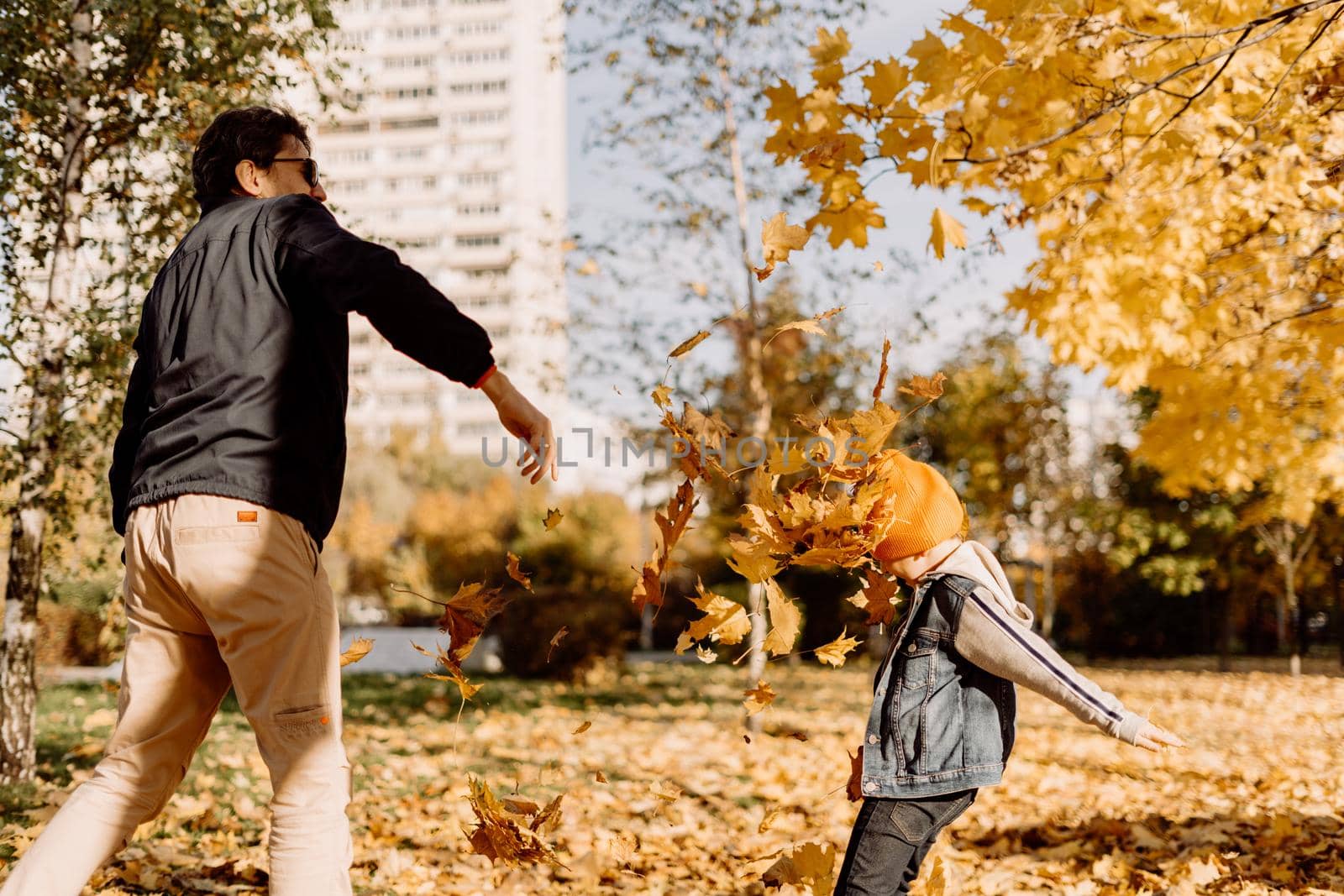 Father and son having fun in autumn park with fallen leaves, throwing up leaf. Child kid boy and his dad outdoors playing with maple leaves. by Ostanina