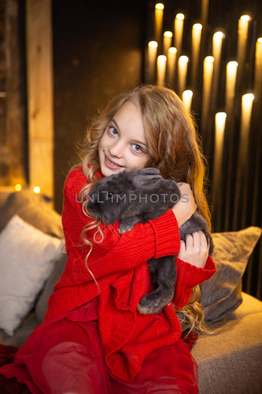 A little blonde girl with a gray rabbit in her arms next to a Christmas tree decorated with garlands, glass balls and Christmas toys and a fireplace. The concept of winter holidays is Christmas and New Year holidays. Magical festive atmosphere.