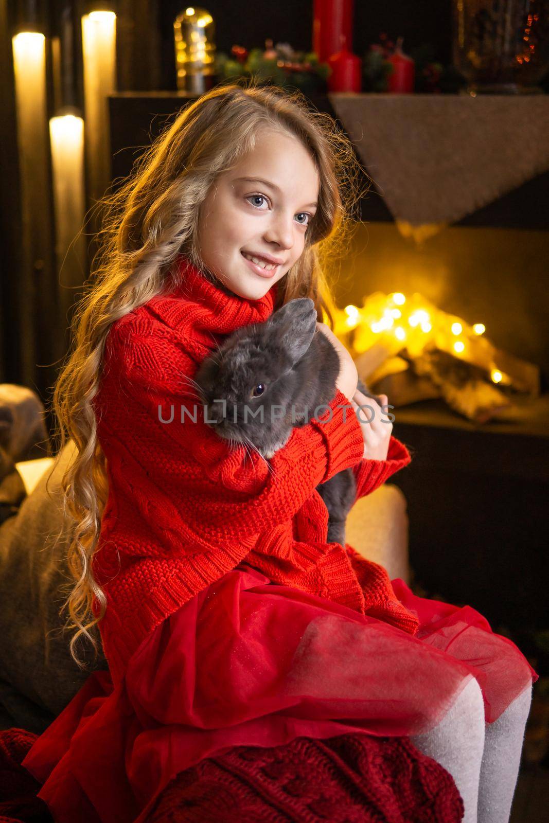 A little blonde girl with a gray rabbit in her arms next to a Christmas tree decorated with garlands by Annu1tochka