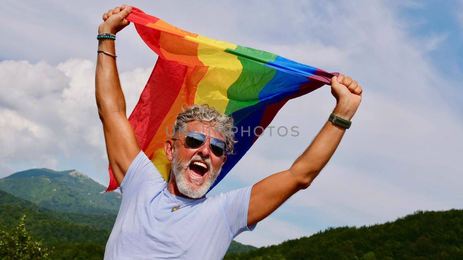 Portrait of a gray-haired elderly Caucasian man with a beard and sunglasses holding a rainbow LGBTQIA flag against a sky background, shouts in protest, Celebrates Pride Month Coming Out Day