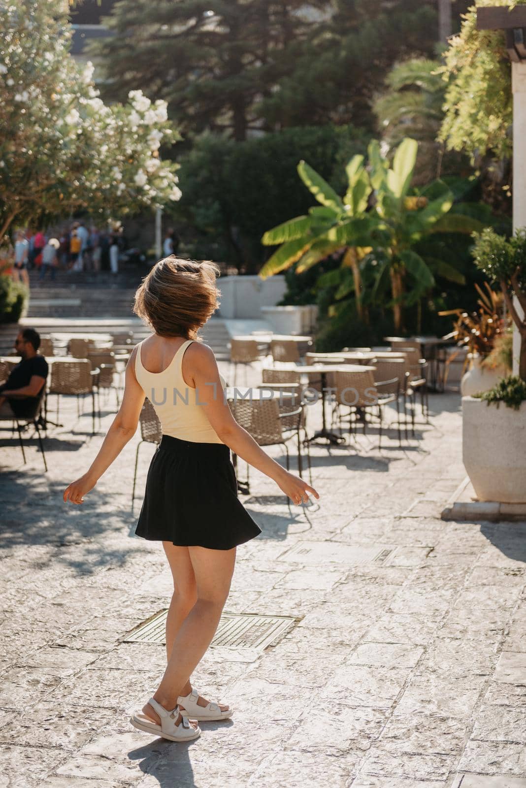 Girl Tourist Walking Through Ancient Narrow Street On A Beautiful Summer Day In MEDITERRANEAN MEDIEVAL CITY, OLD TOWN BUDVA, MONTENEGRO. Young Beautiful Cheerful Woman Walking On Old Street At Tropical Town. Pretty Girl Looking At You And Smiling by Andrii_Ko