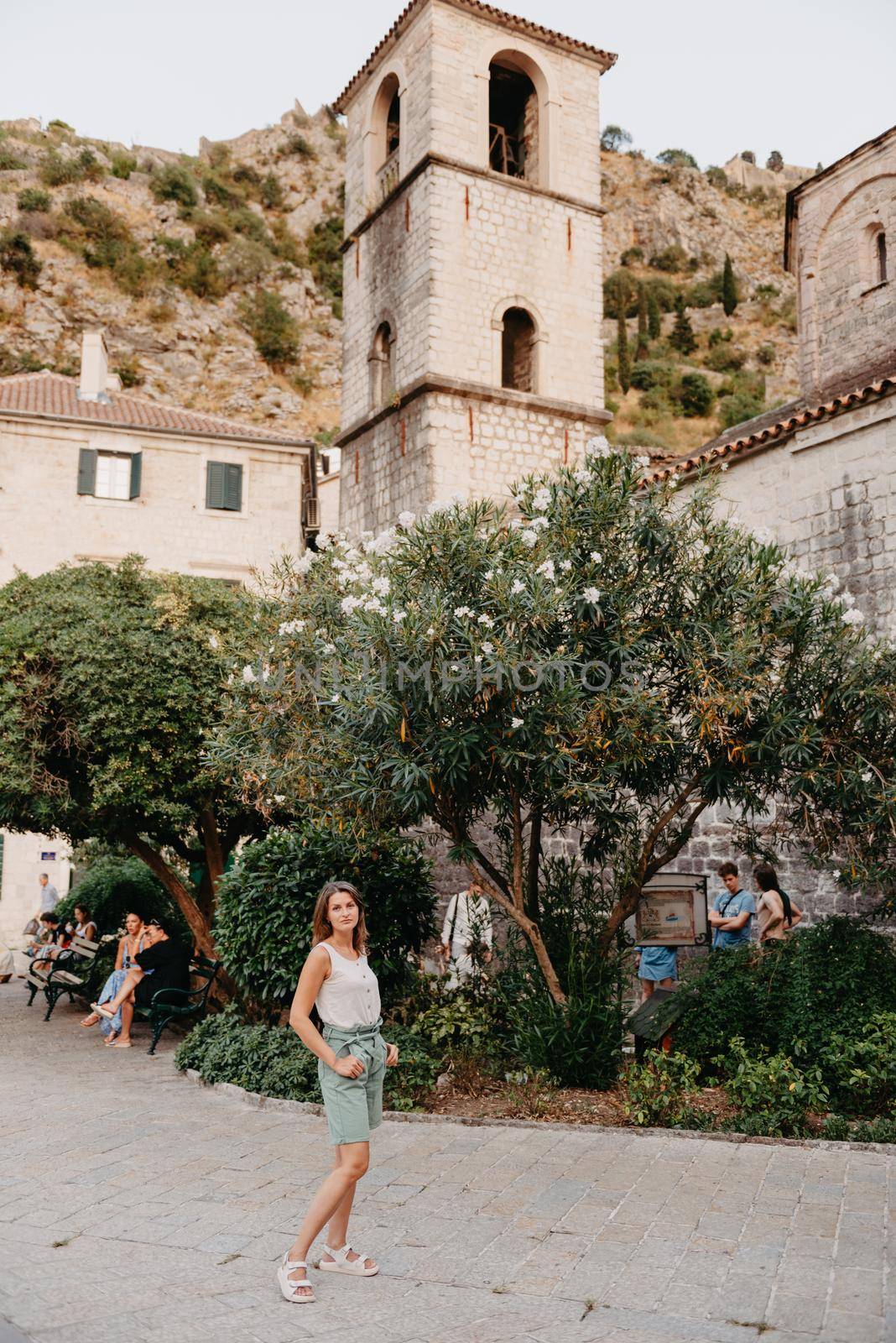 Girl Tourist Resting in the Ancient Narrow Street On A Beautiful Summer Day In MEDITERRANEAN MEDIEVAL CITY, OLD TOWN KOTOR, MONTENEGRO. Young Beautiful Cheerful Woman Walking On Old Street