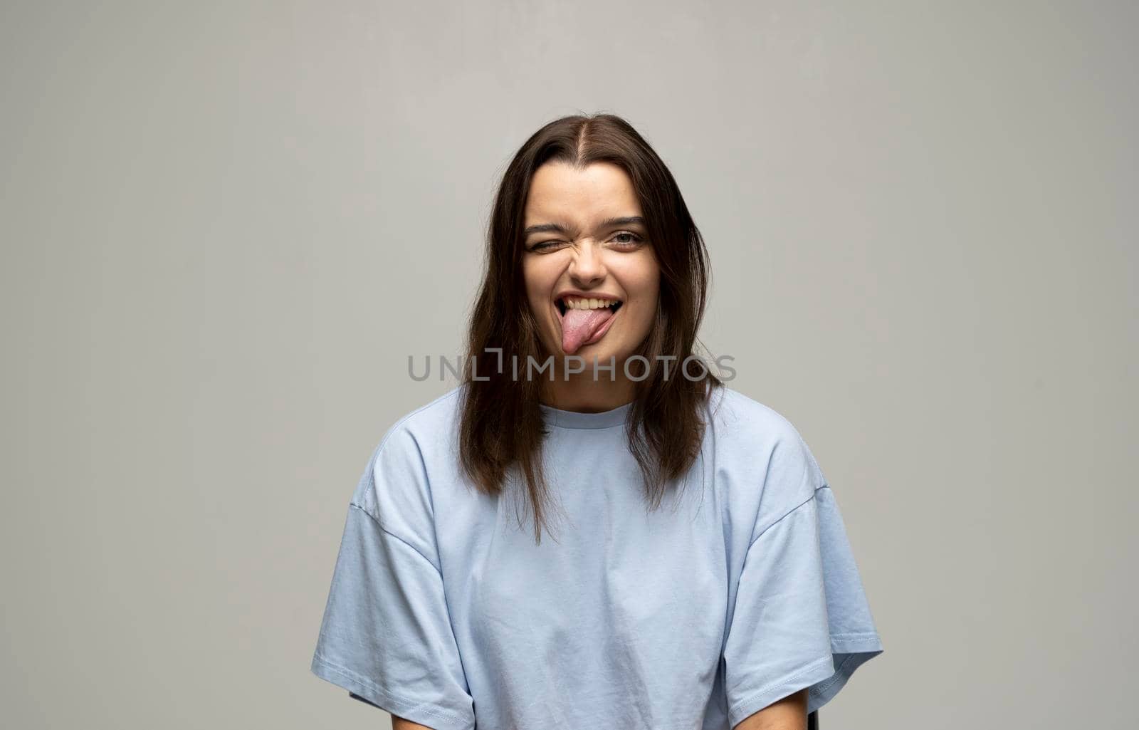 Portrait of young beautiful cute cheerful brunette girl in blue t-shirt smiling looking at camera and showing a tongue over white background studio. Emotions