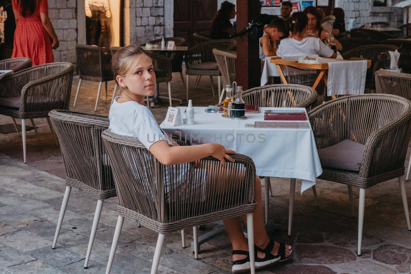 Girl Tourist Resting in the Ancient Narrow Street On A Beautiful Summer Day In MEDITERRANEAN MEDIEVAL CITY, OLD TOWN KOTOR, MONTENEGRO. Young Beautiful Cheerful Woman Walking On Old Street