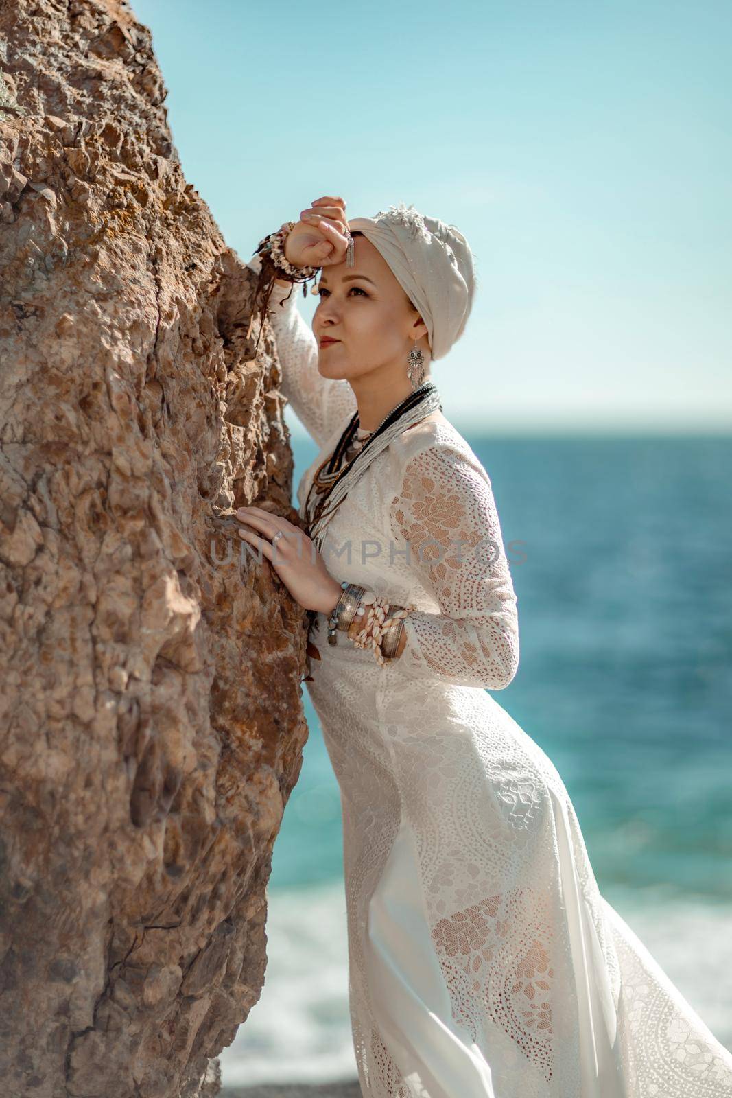 Middle aged woman looks good with blond hair, boho style in white long dress on the beach decorations on her neck and arms. by Matiunina