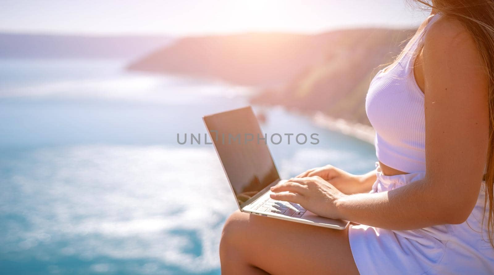 Successful business woman in yellow hat working on laptop by the sea. Pretty lady typing on computer at summer day outdoors. Freelance, travel and holidays concept.