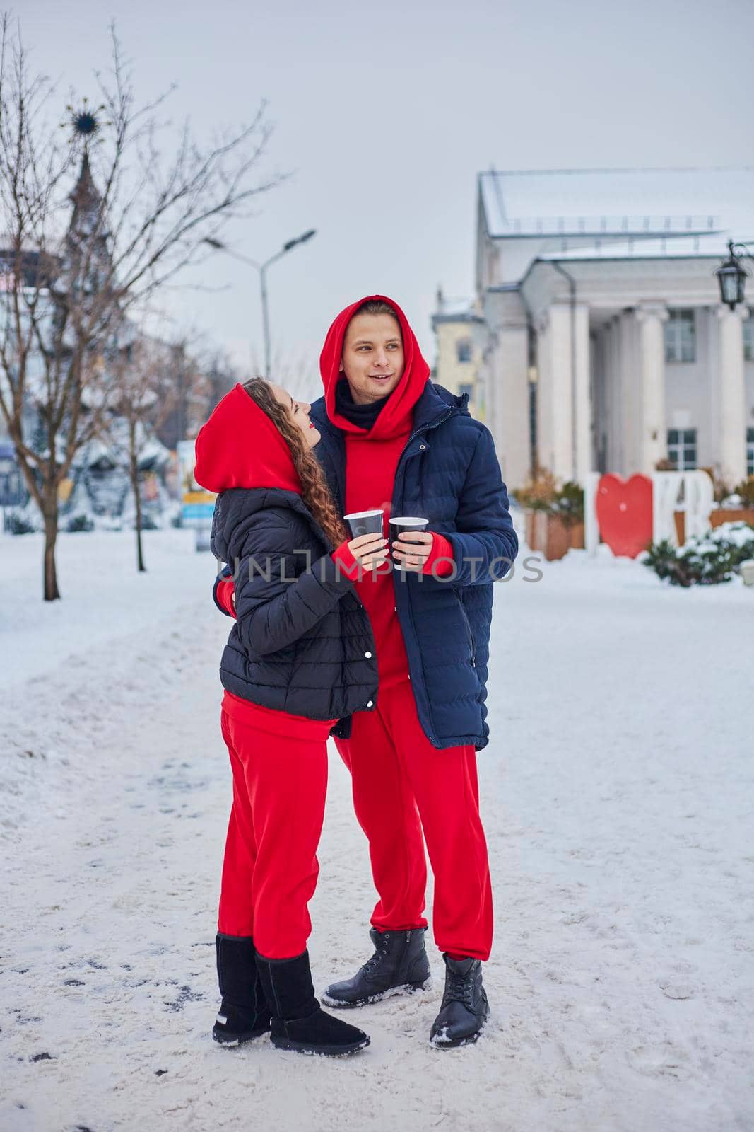 young family guy and girl spend the day in the park on a snowy day. the guy hugs the girl while standing on the street, they drink coffee together