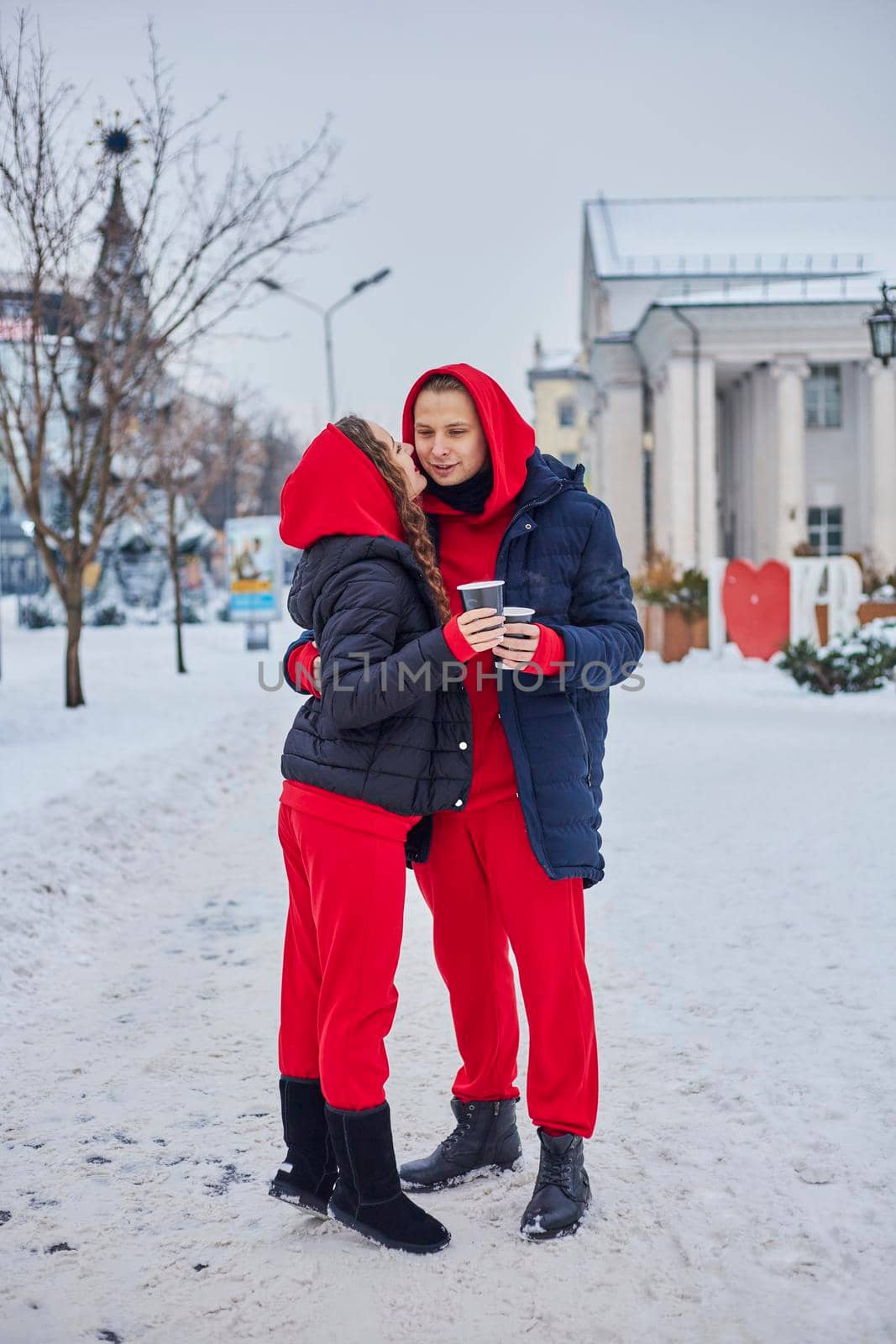 young family guy and girl spend the day in the park on a snowy day. the guy hugs the girl while standing on the street, they drink coffee together