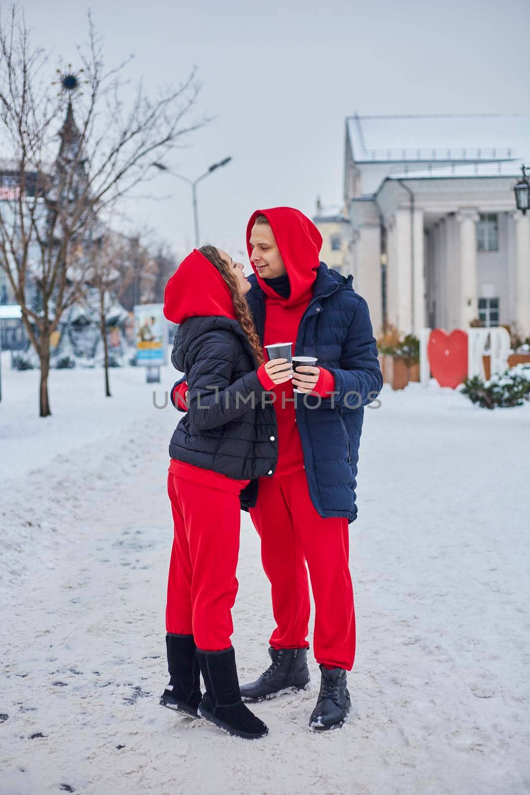 young family guy and girl spend the day in the park on a snowy day. the guy hugs the girl while standing on the street, they drink coffee together. by mosfet_ua