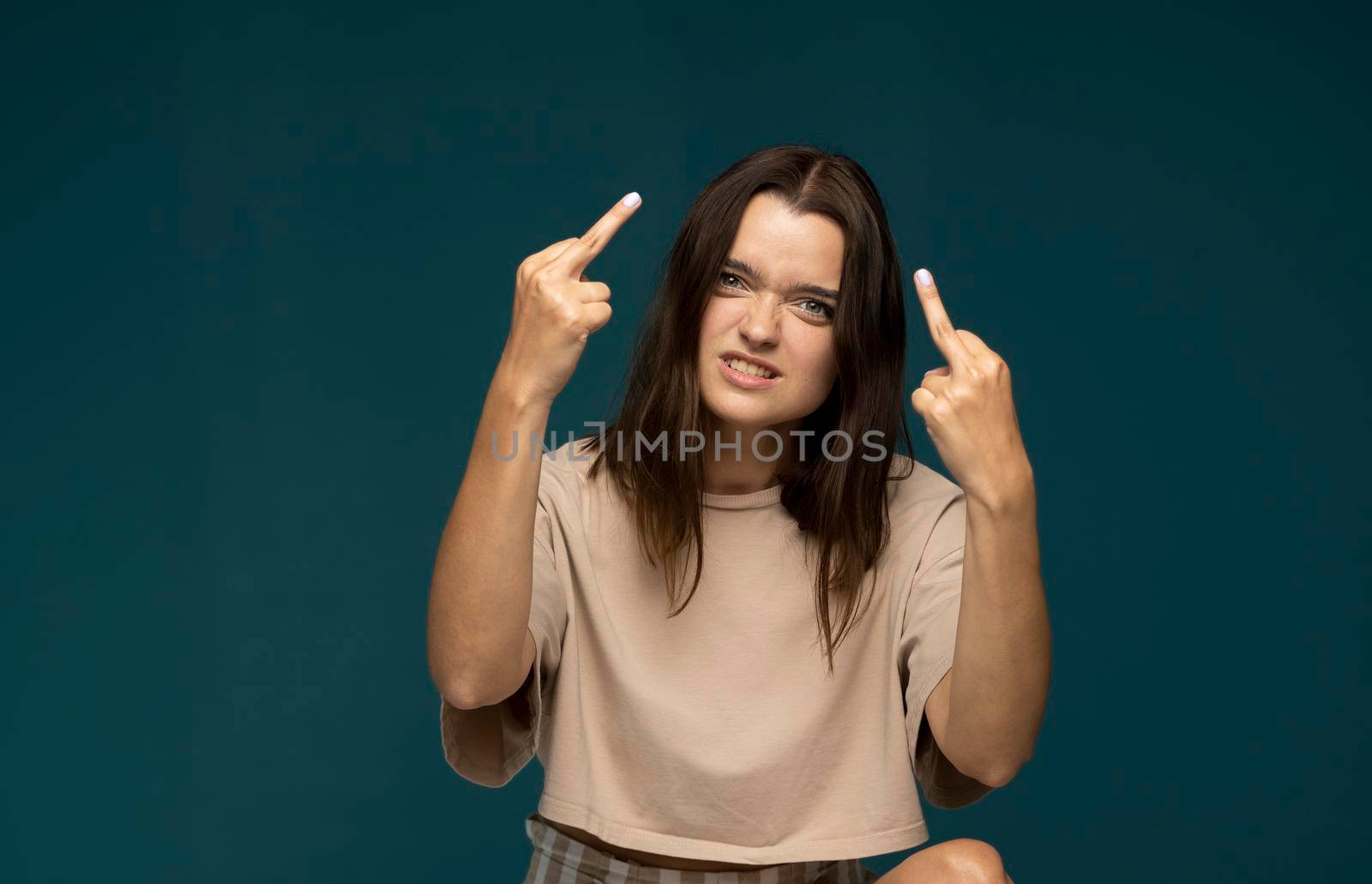 Portrait of brunette student woman showing middle finger doing rebel expression at camera standing in studio with blue background