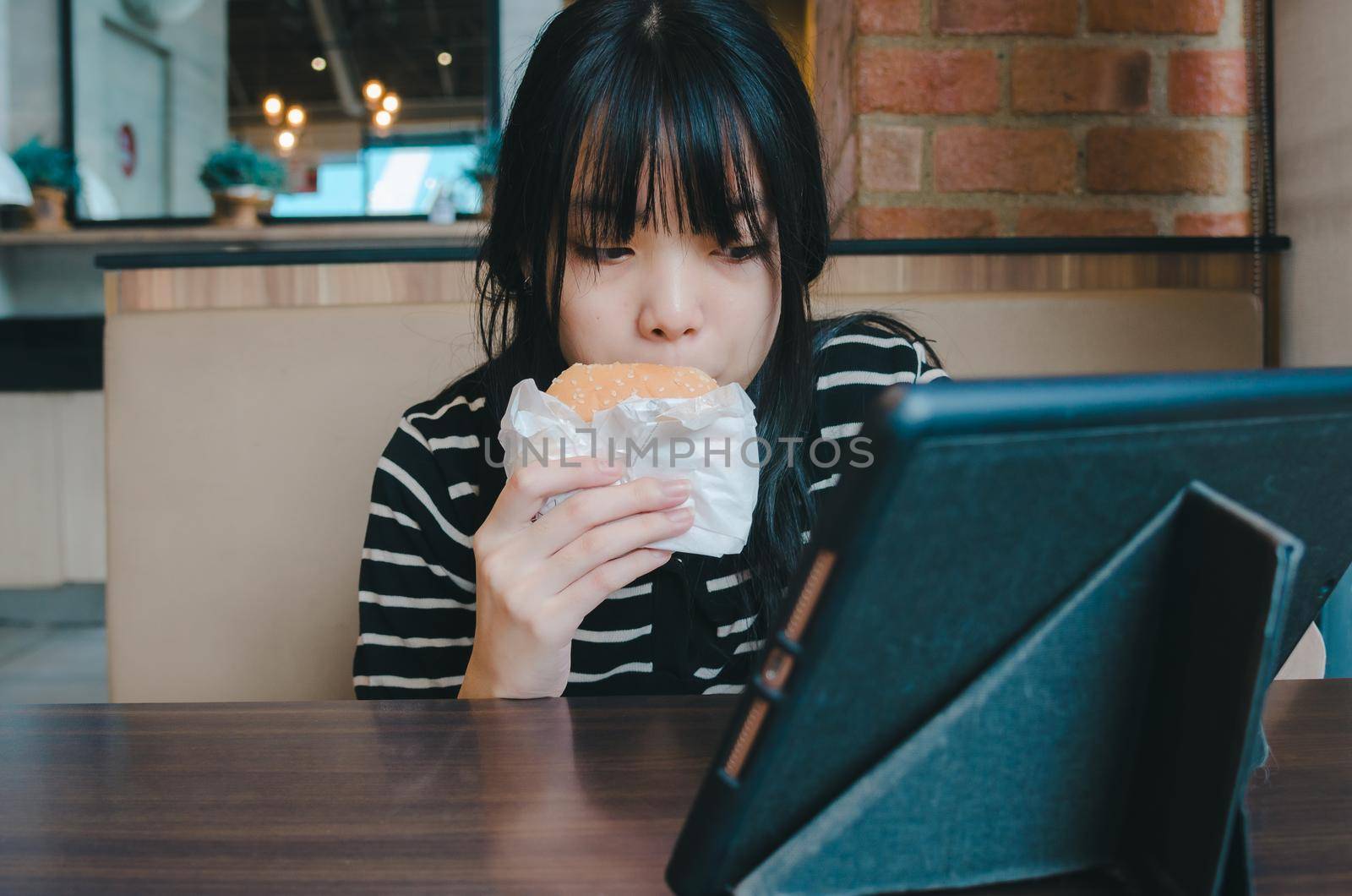 young girl is eating a hamburger and watching an online social media tech tablet on the table.