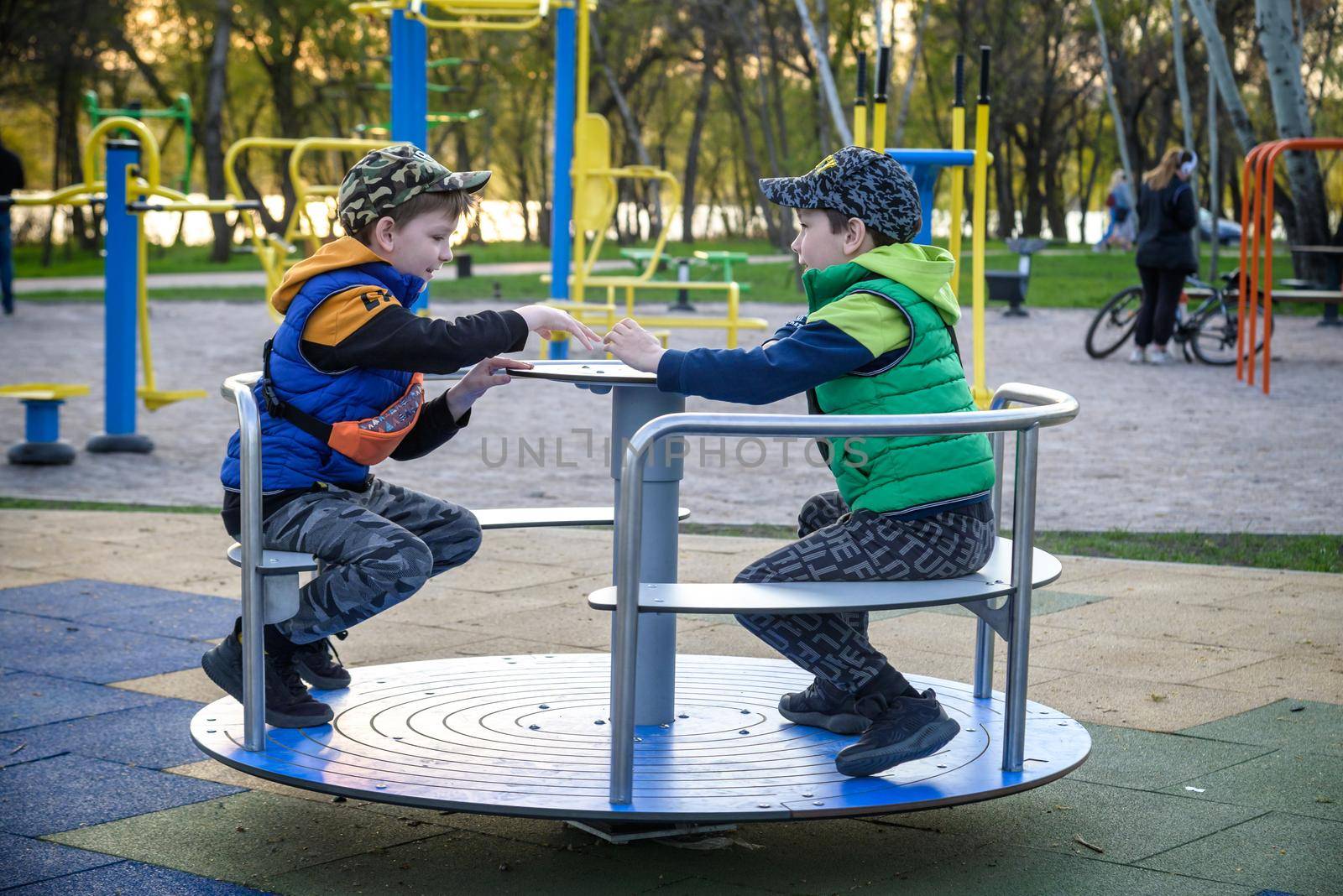 Brightly dressed twin brothers play on a carousel in an autumn park amid fallen yellow leaves. They are two years old.
