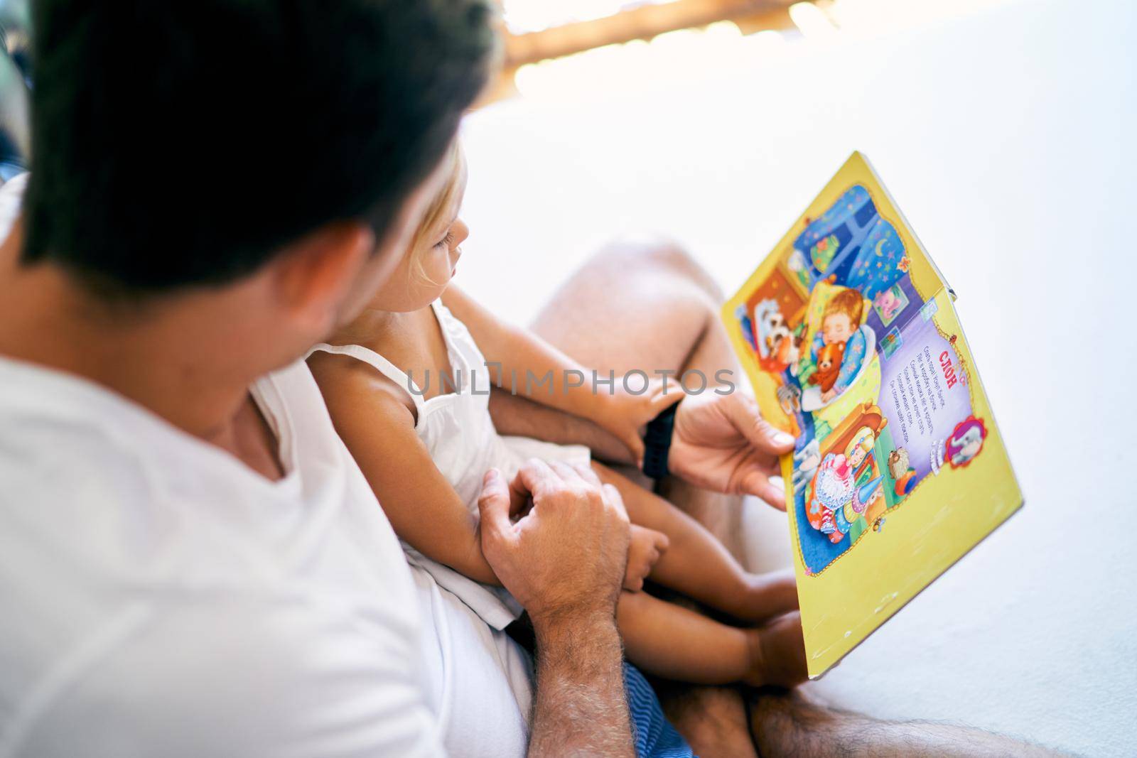 Dad reads a book of fairy tales to a little girl on the bed. High quality photo