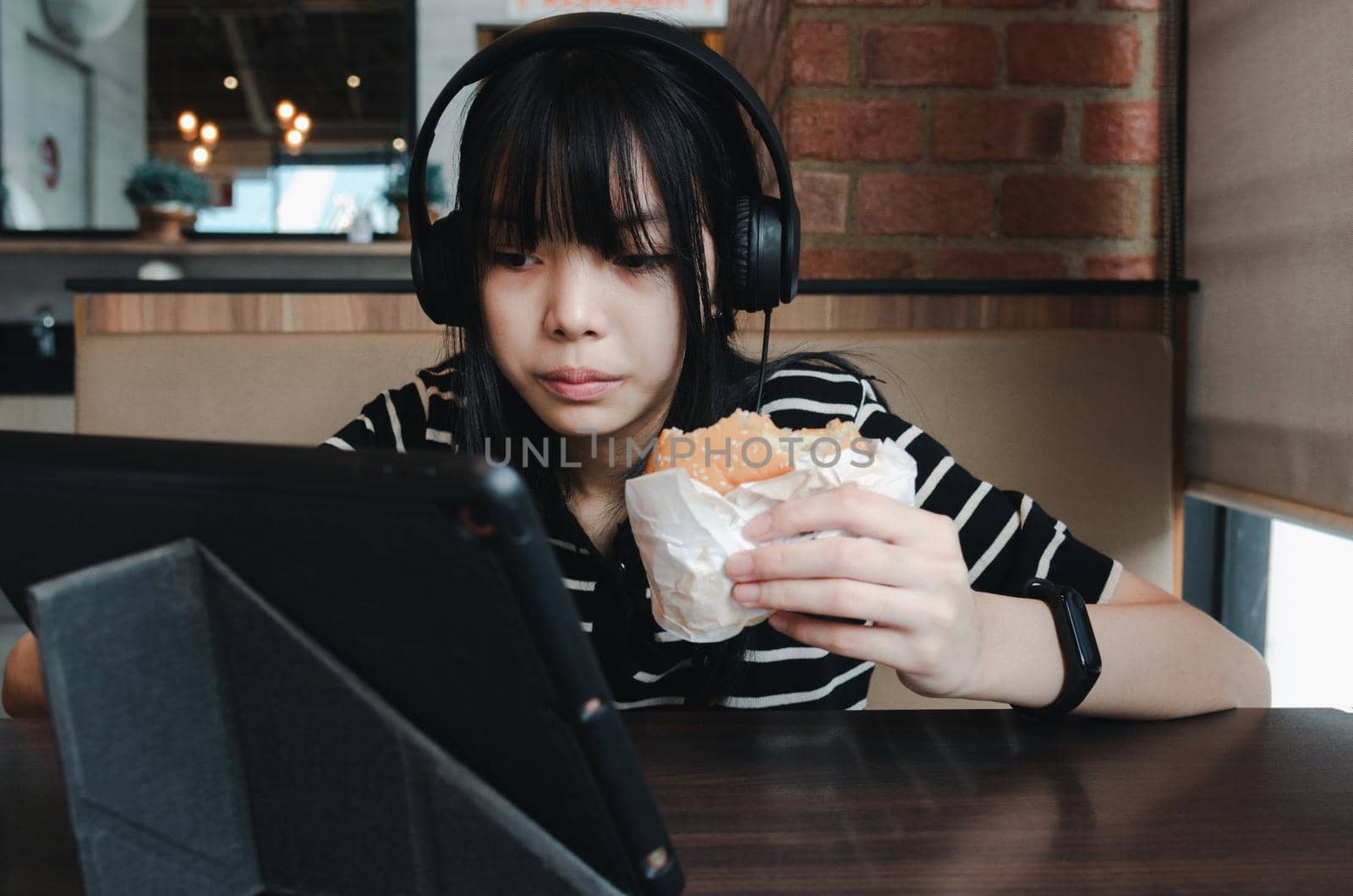 Girl eating burger watch your tablet and use headphones to listen to music or use social media.