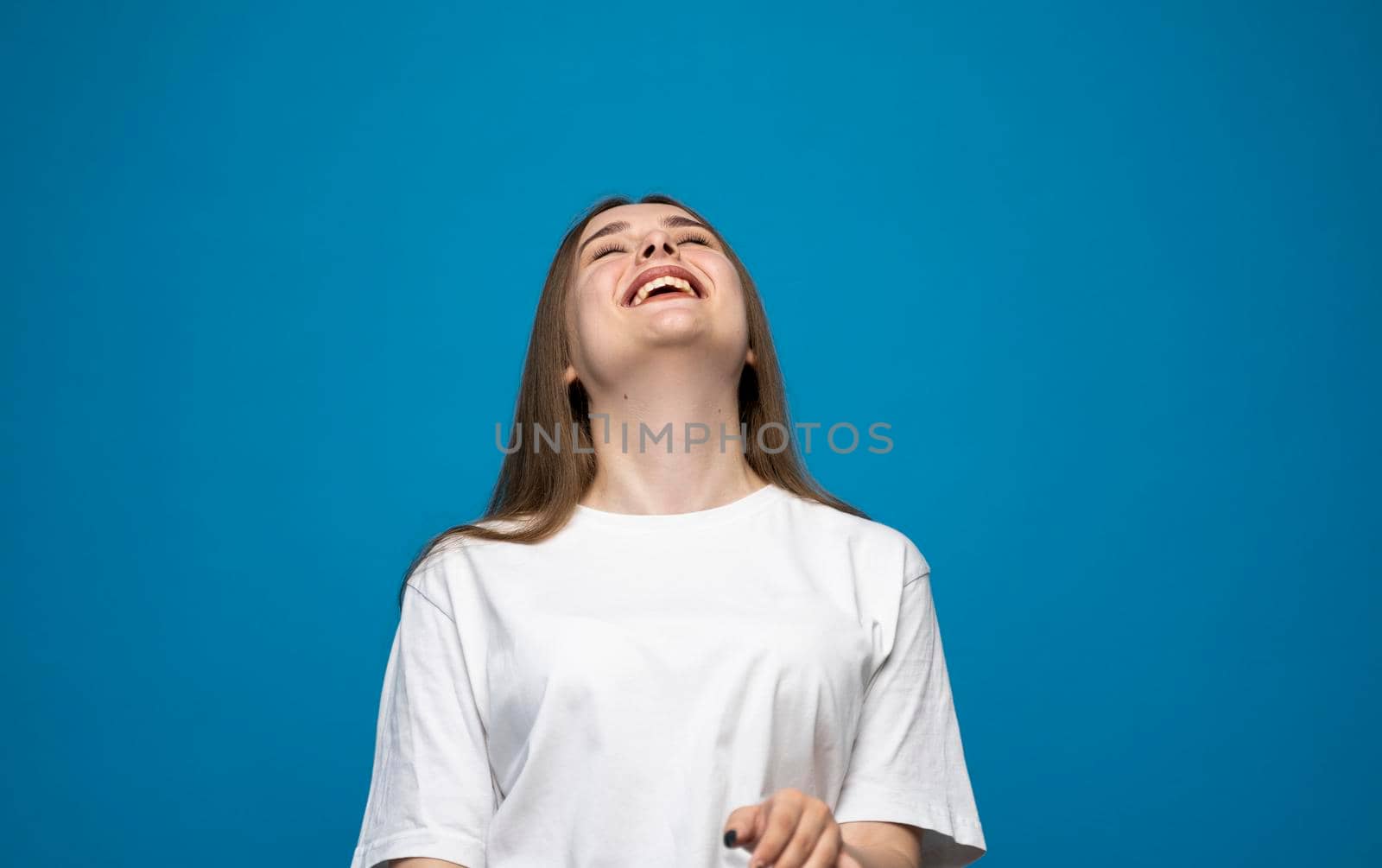 Young beautiful cute cheerful brunette girl in white t-shirt smiling looking at camera and showing a tongue over blue background studio. Emotions