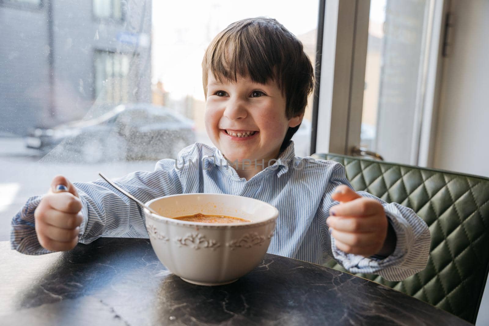 Boy laughing at the cafe while sitting at the window with a big bowl of soup. High quality photo