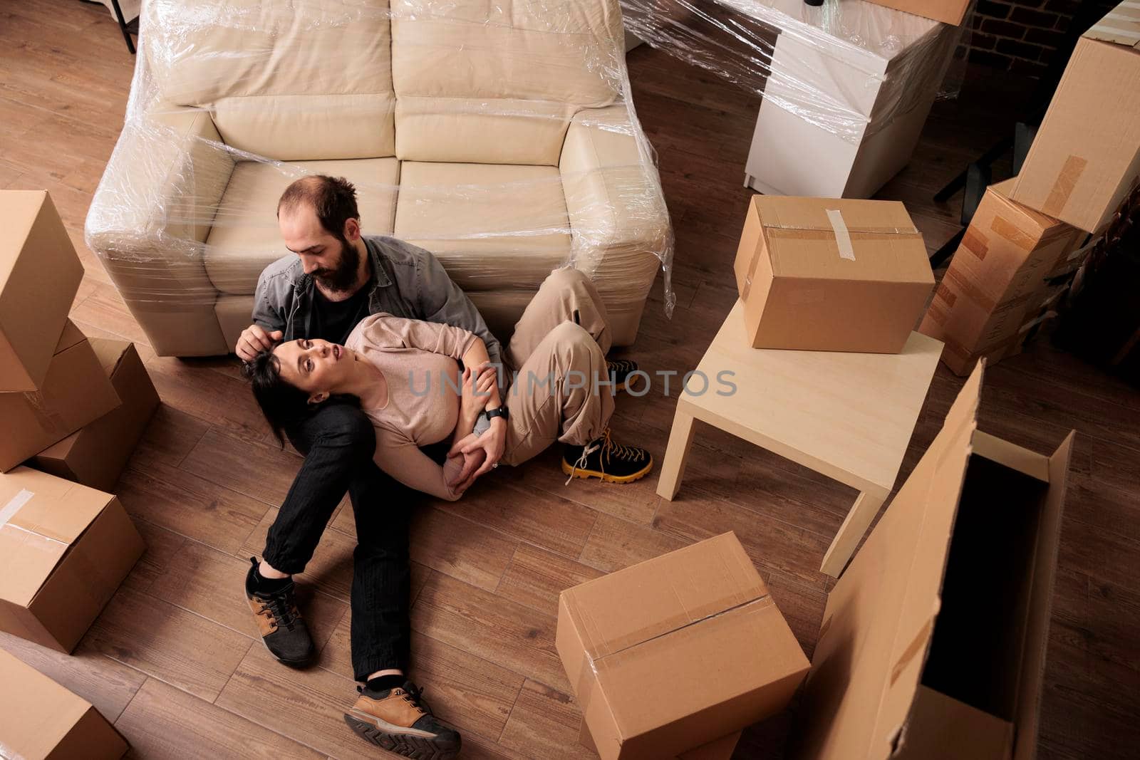 Happy man and woman sitting on living room floor together, relaxing after relocating in new rented apartment household. Moving in with furniture and decorations, relationship event. Top view of.