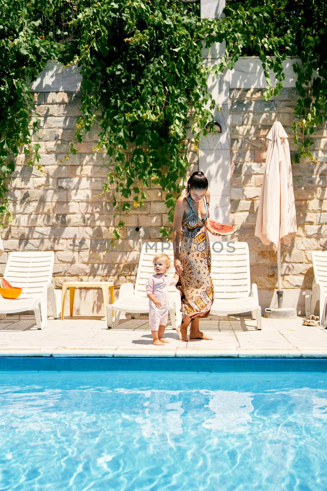 Mom with a watermelon in her hand leads a little girl to a sun lounger by the pool. High quality photo