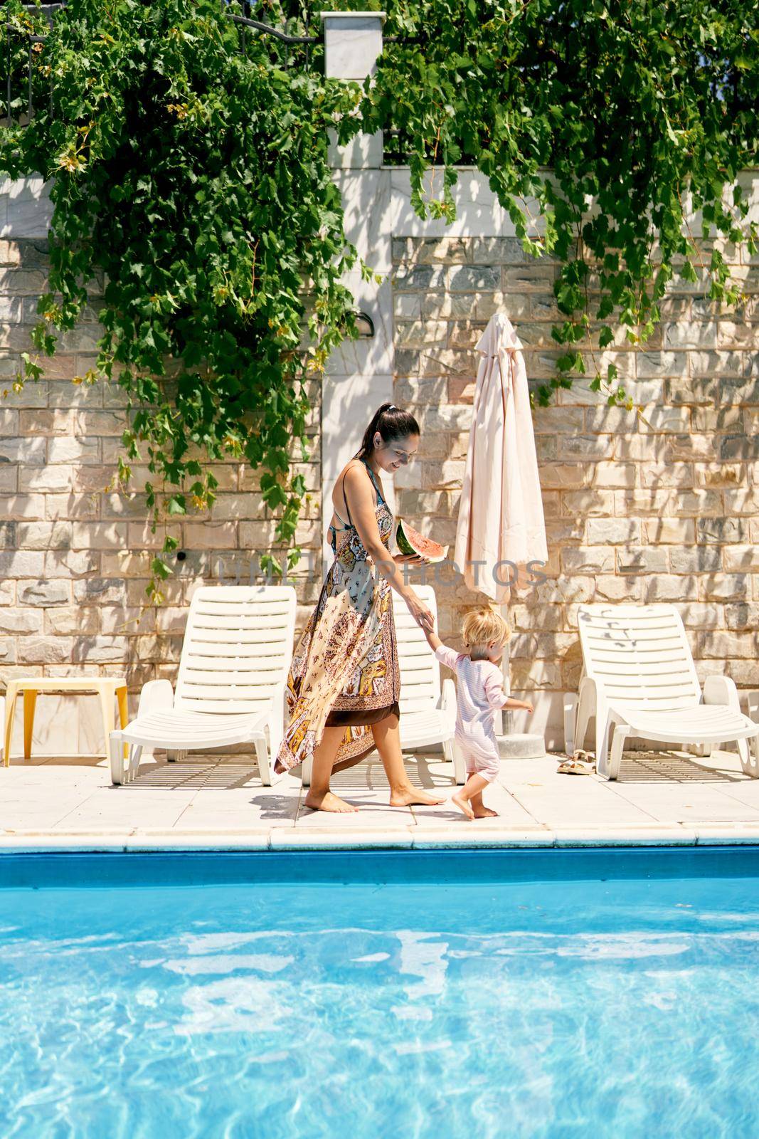 Mom with a watermelon in her hand leads a little girl to a sun lounger. High quality photo