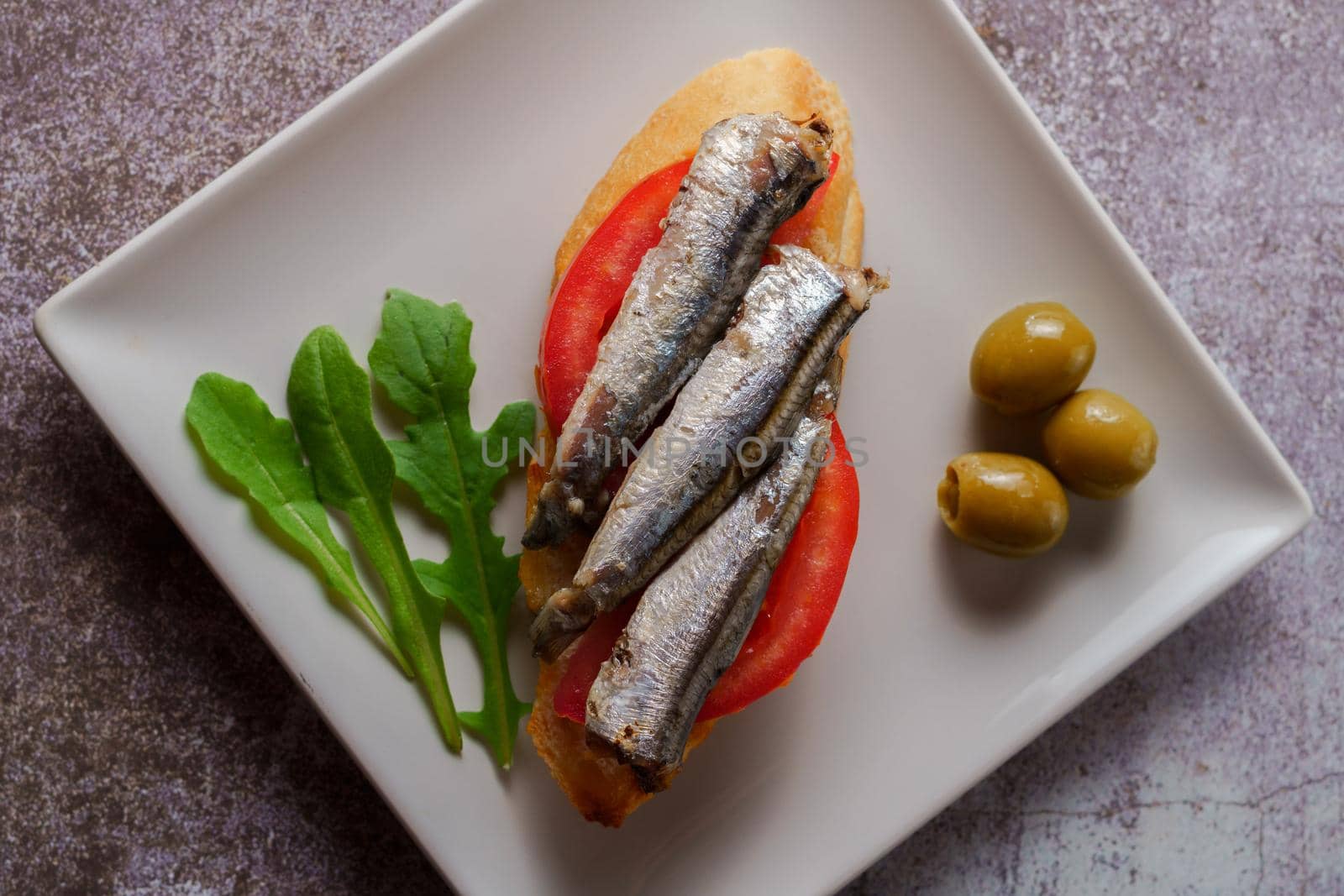 tapa of sardines on a slice of bread with tomato and olives on a white plate with a typical spanish white background