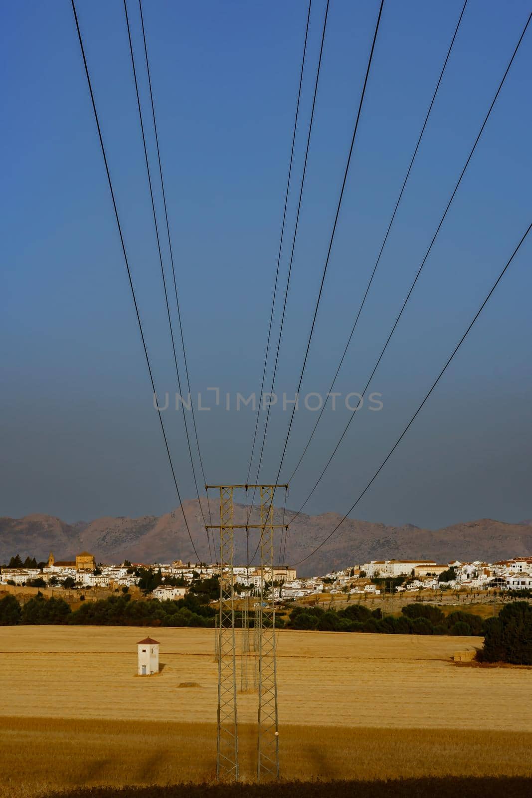 electric tower with cables, in the background a white village with cereal fields.