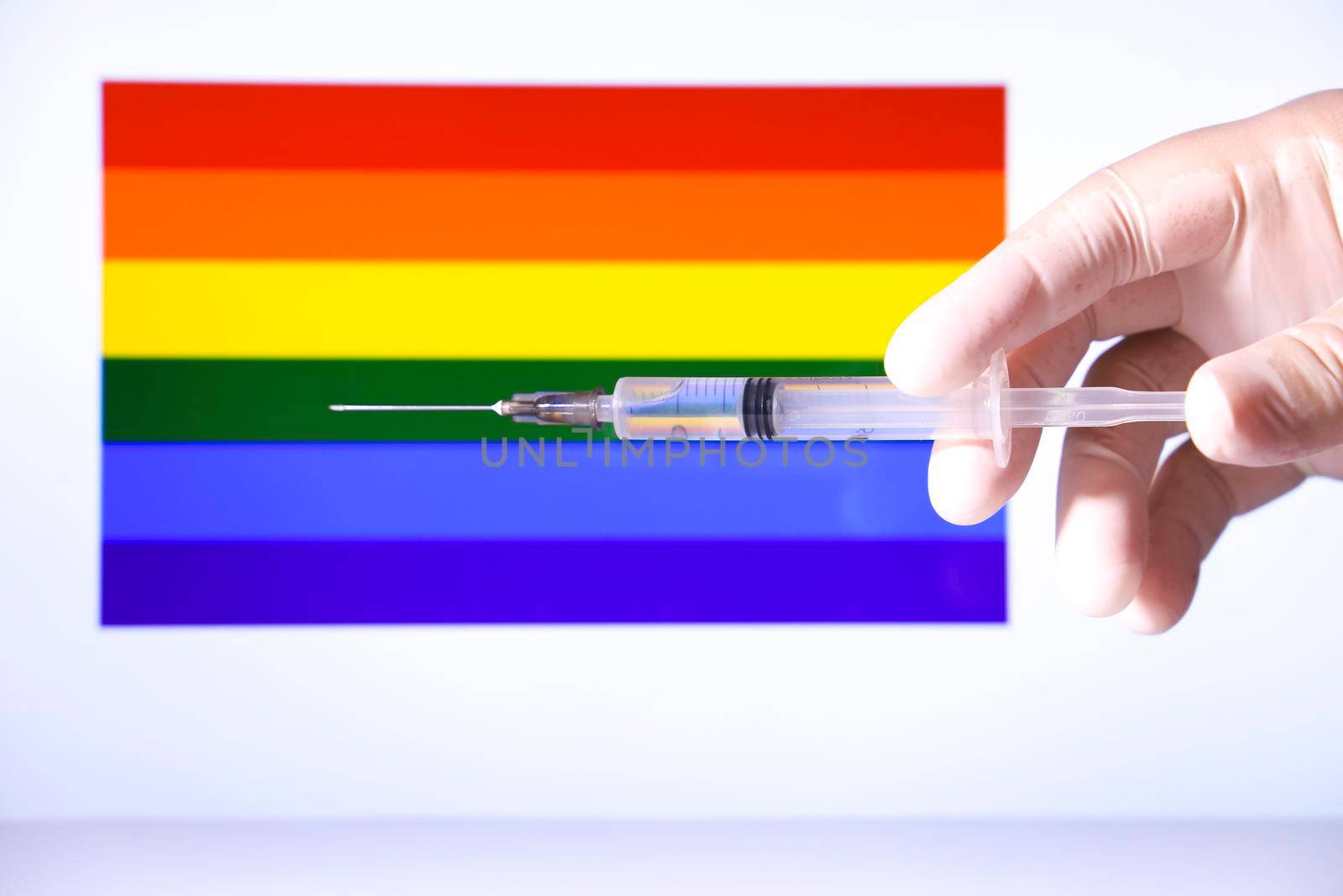 Hand in surgical glove holding syringe and rainbow flag in the background
