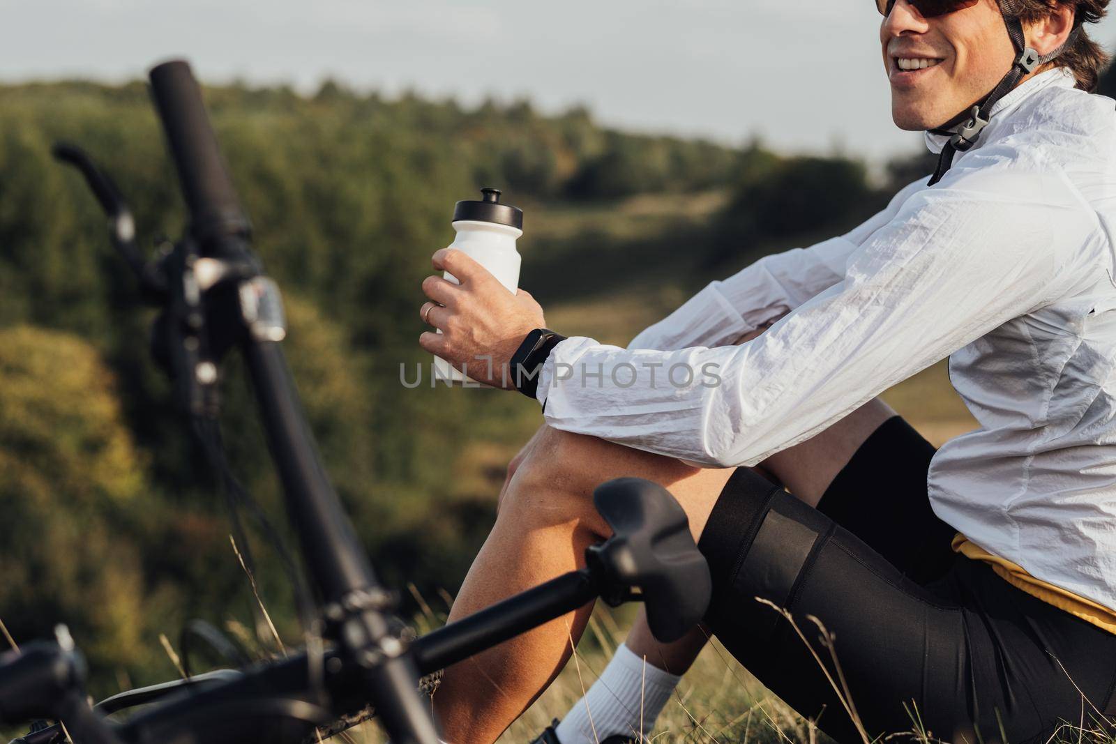 Professional Male Cyclist Drinking Water from Bottle, Man Sitting Near Bicycle During His Journey Outdoors in Countryside