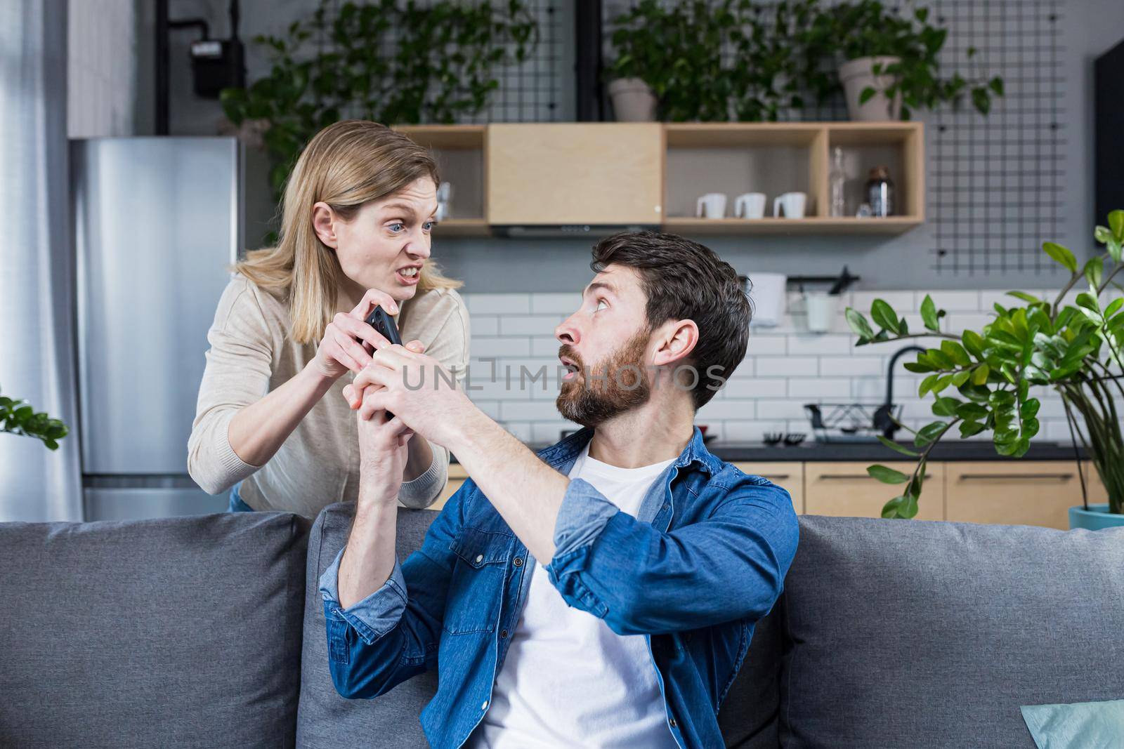Conflict in the family. An angry, annoyed woman's wife takes the phone from the man who is sitting on the couch at home and constantly uses the phone.