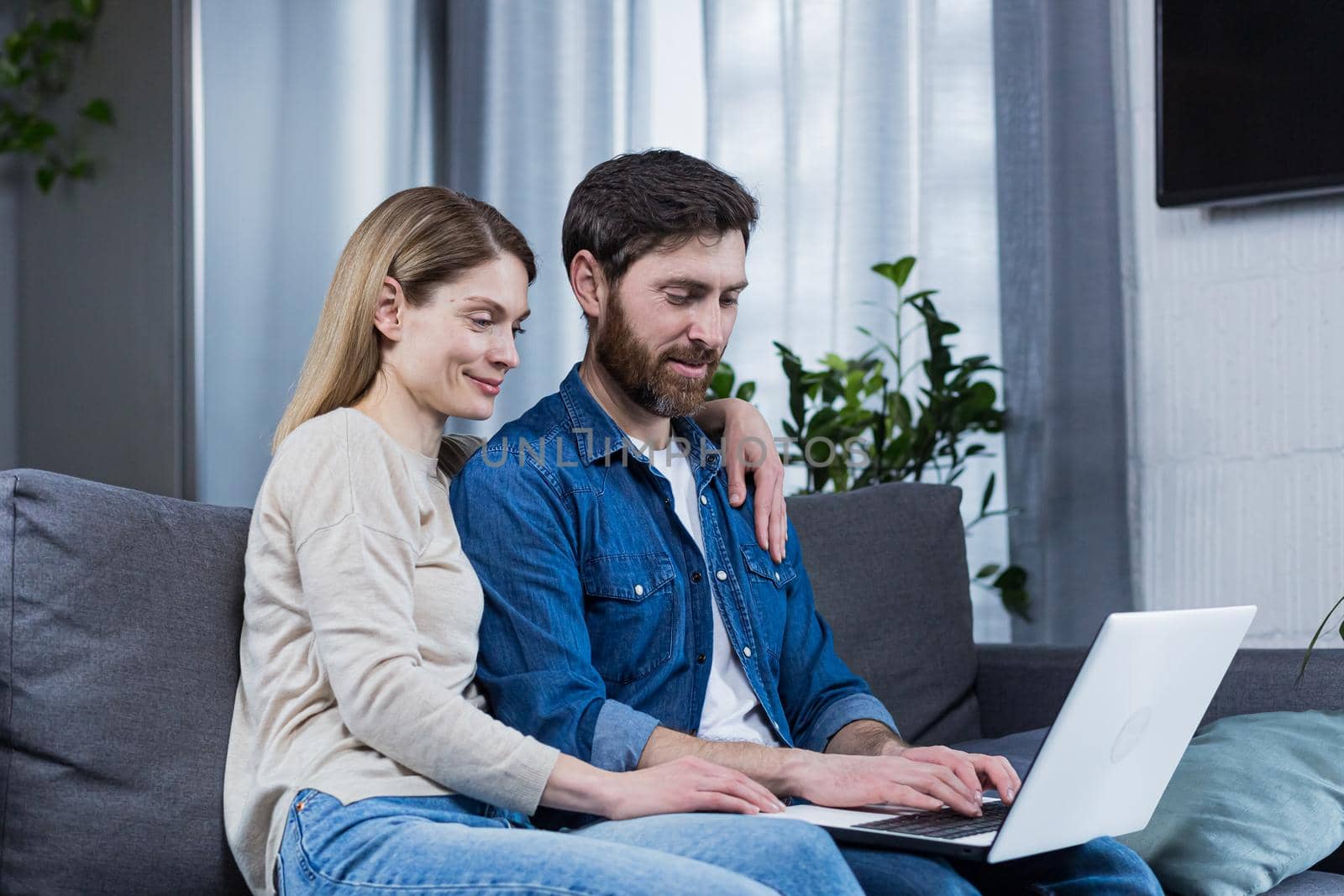 Happy young married couple man and woman together at home, sitting on sofa smiling and using laptop