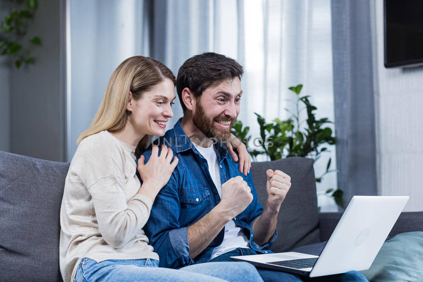 Happy couple man and woman looking at laptop screen, celebrating victory, holding hands up, gesture of victory and success