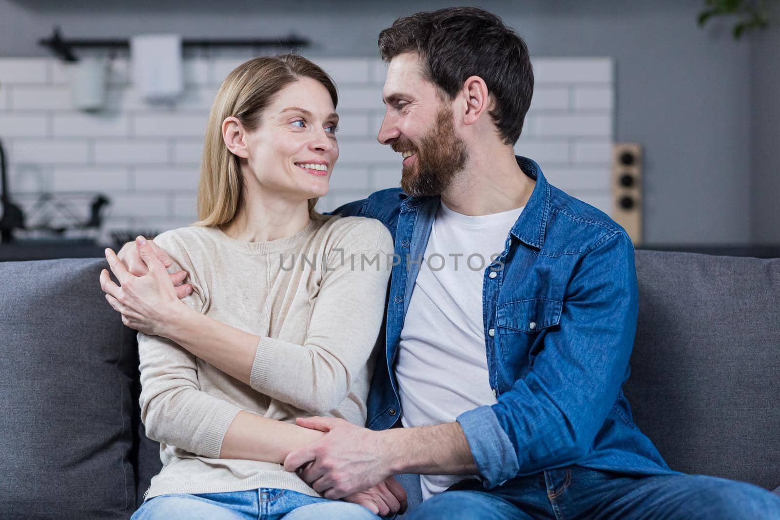 Portrait of a happy young family, couple. Man and woman sitting on sofa, hugging, waving, smiling, looking at camera, looking at each other