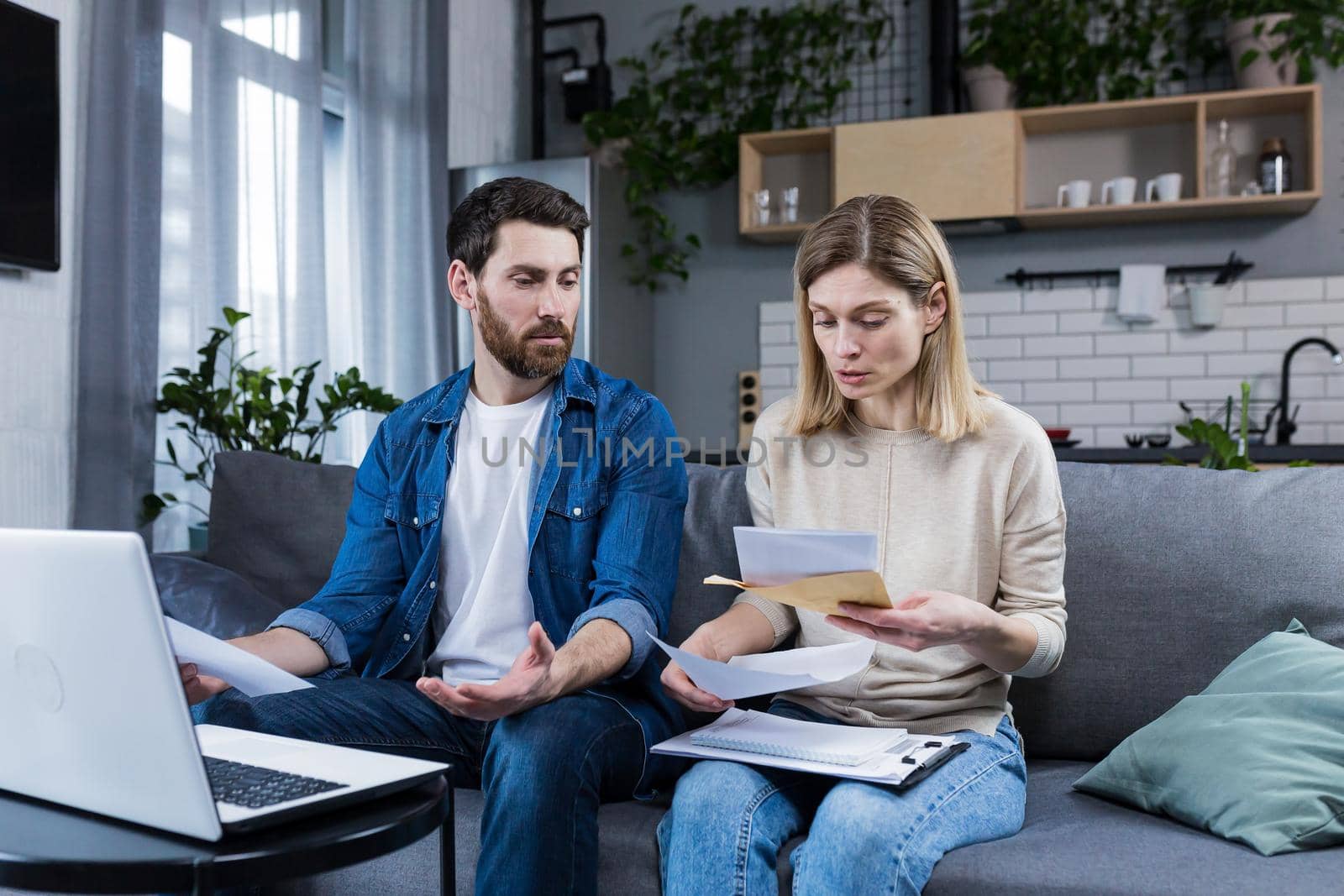 Young family, husband and wife, reviewing their bills, loan and mortgage agreements, sitting on the couch at home, working with documents on a laptop, embarrassed, upset