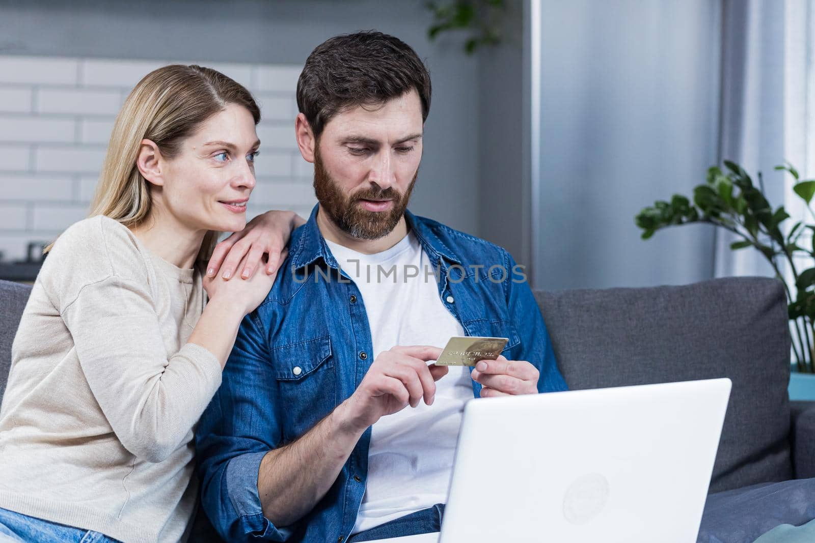 Happy married couple man and woman sitting at home on the couch using laptop for online shopping, holding a credit bank card
