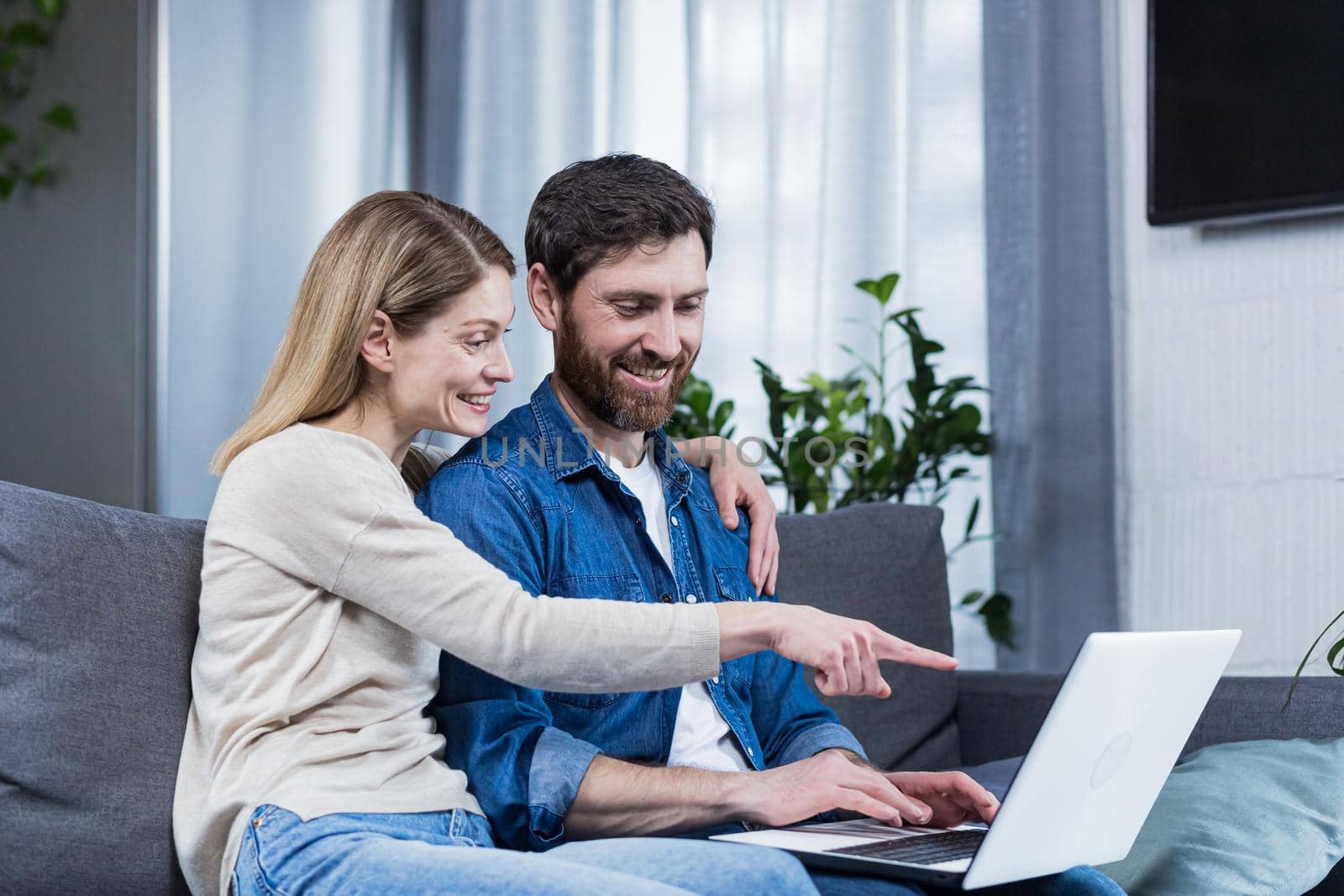 Young couple, family, husband and wife sitting on sofa at home, using laptop, winning something, getting good news, rejoicing, smiling, hugging