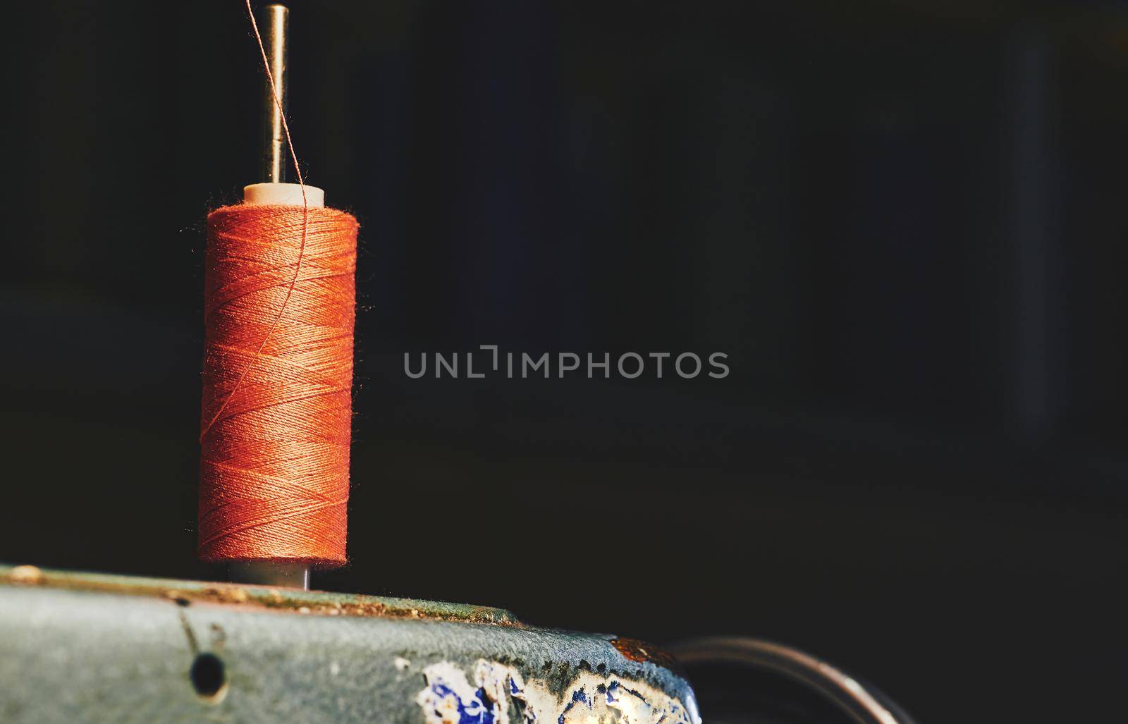a long, thin strand of cotton, nylon, or other fibers used in sewing or weaving. Orange spool of thread on an old gray sewing machine and a dark background.