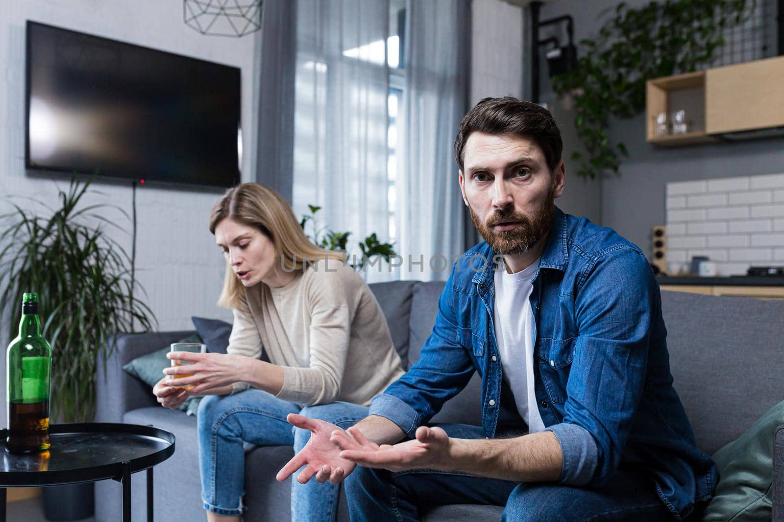 Female alcoholism, husband in despair, woman wife drinking hard alcohol, couple sitting on sofa in living room