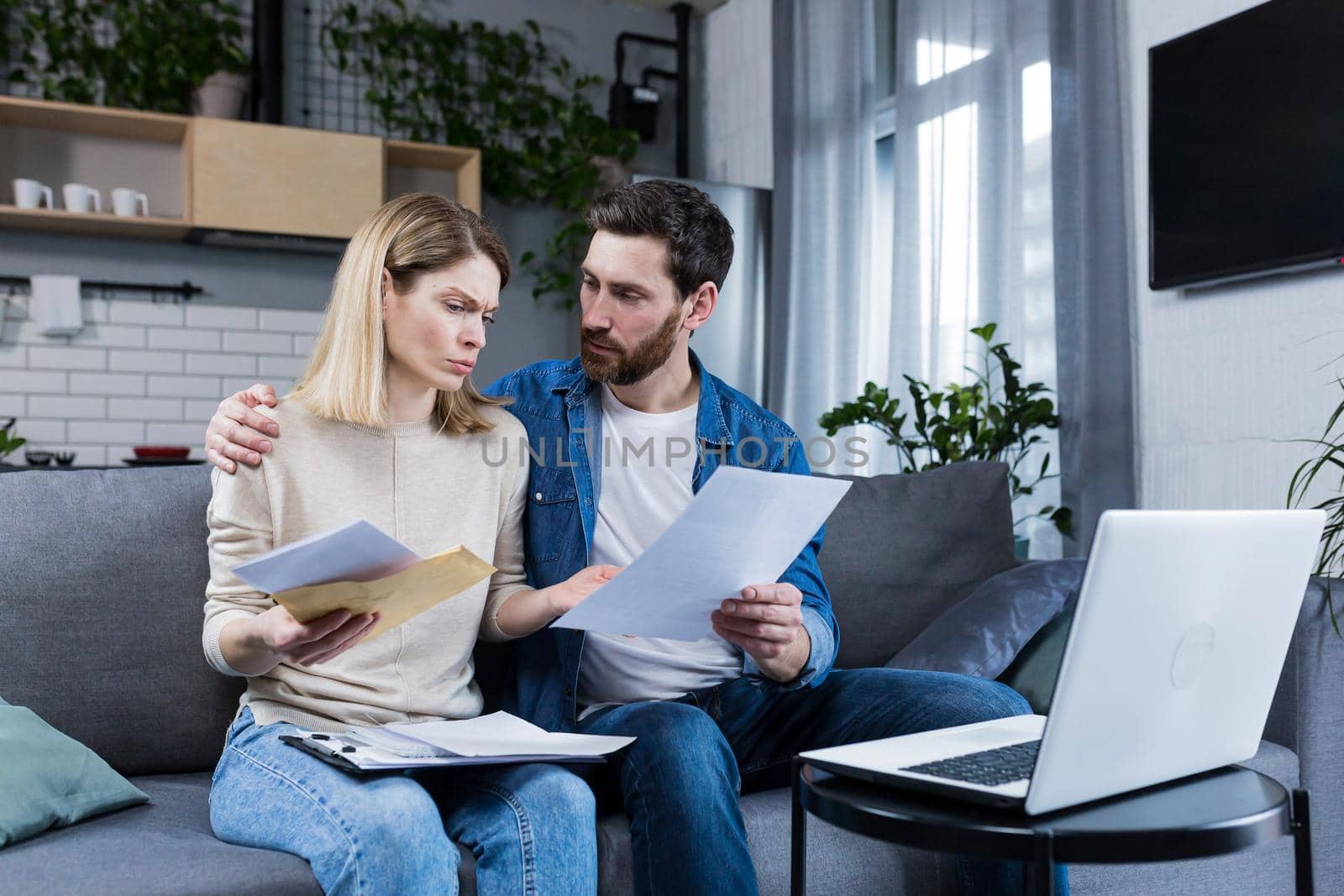 Young family, husband and wife, reviewing their bills, loan and mortgage agreements, sitting on the couch at home, working with documents on a laptop, embarrassed, upset