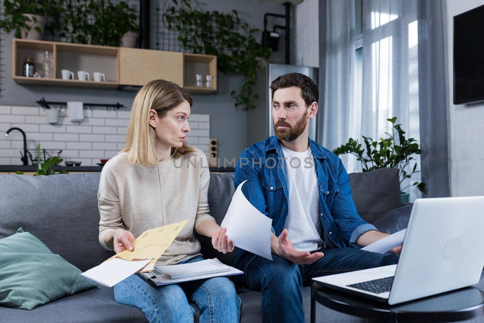 Young family, husband and wife, reviewing their bills, loan and mortgage agreements, sitting on the couch at home, working with documents on a laptop, embarrassed, upset