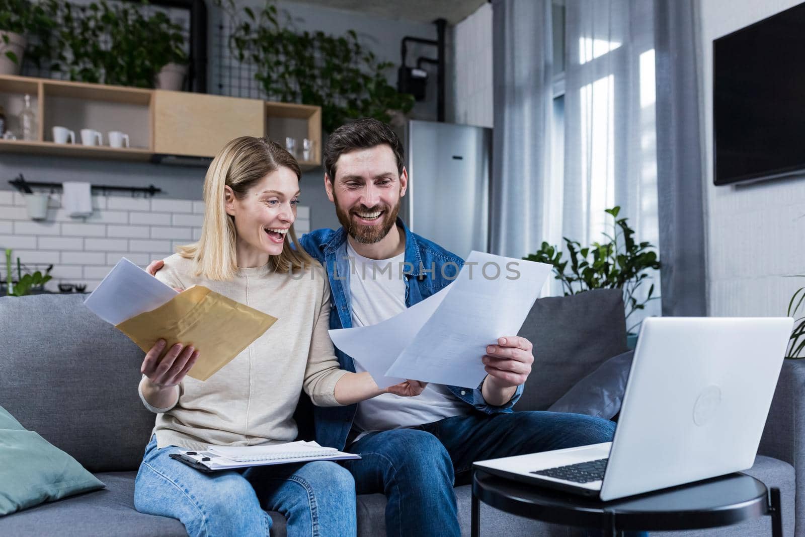 Happy married couple, man and woman, received a positive letter, sitting at home on the couch, smiling and rejoicing