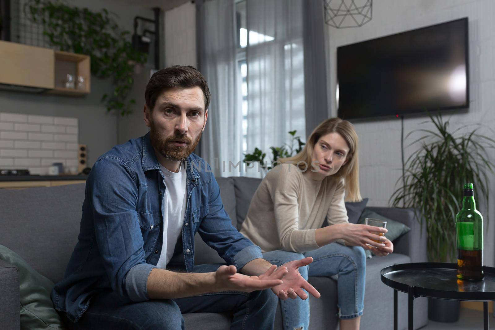 Female alcoholism, husband in despair, woman wife drinking hard alcohol, couple sitting on sofa in living room