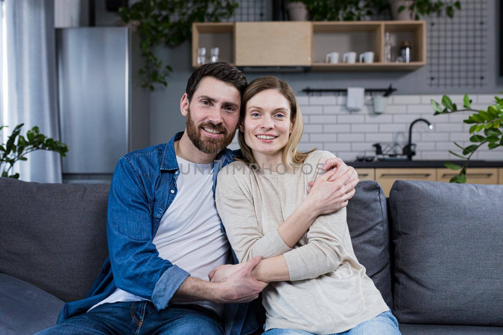 Portrait of a happy young family, couple. Man and woman sitting on sofa, hugging, waving, smiling, looking at camera, looking at each other